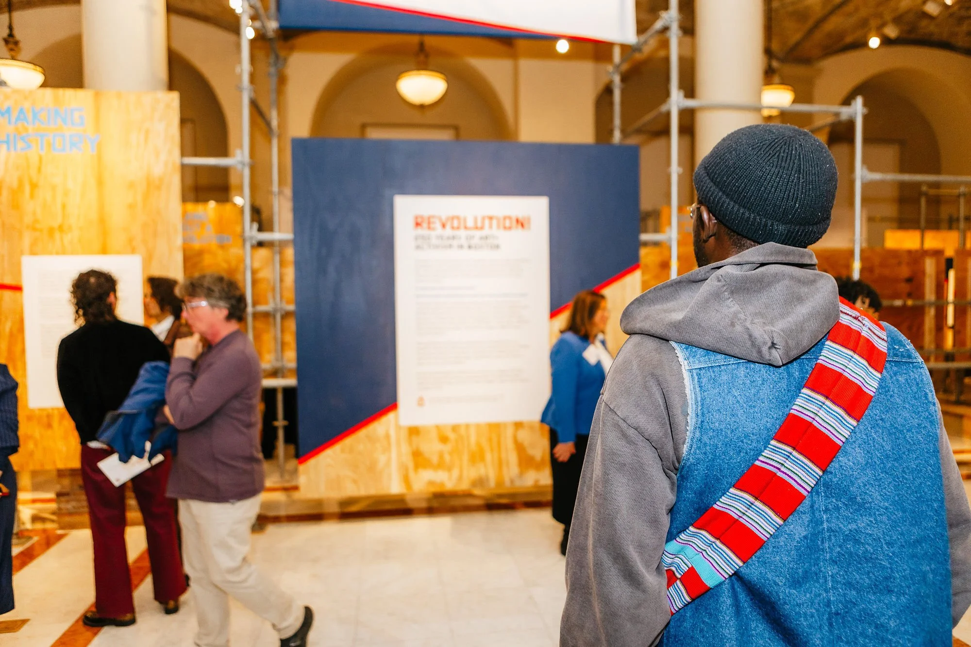A man in a gray hoodie and black knit cap stands in a museum exhibition space, observing display boards. Several people are visible in the background, engaging with educational displays about history and revolution, with the space featuring high ceilings, large columns, and warm lighting.