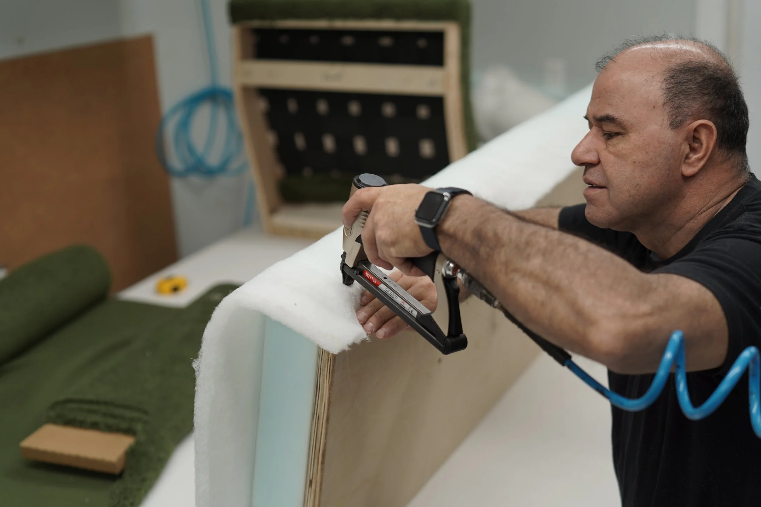 A man assembling or upholstering furniture using a stapler gun in a workshop.