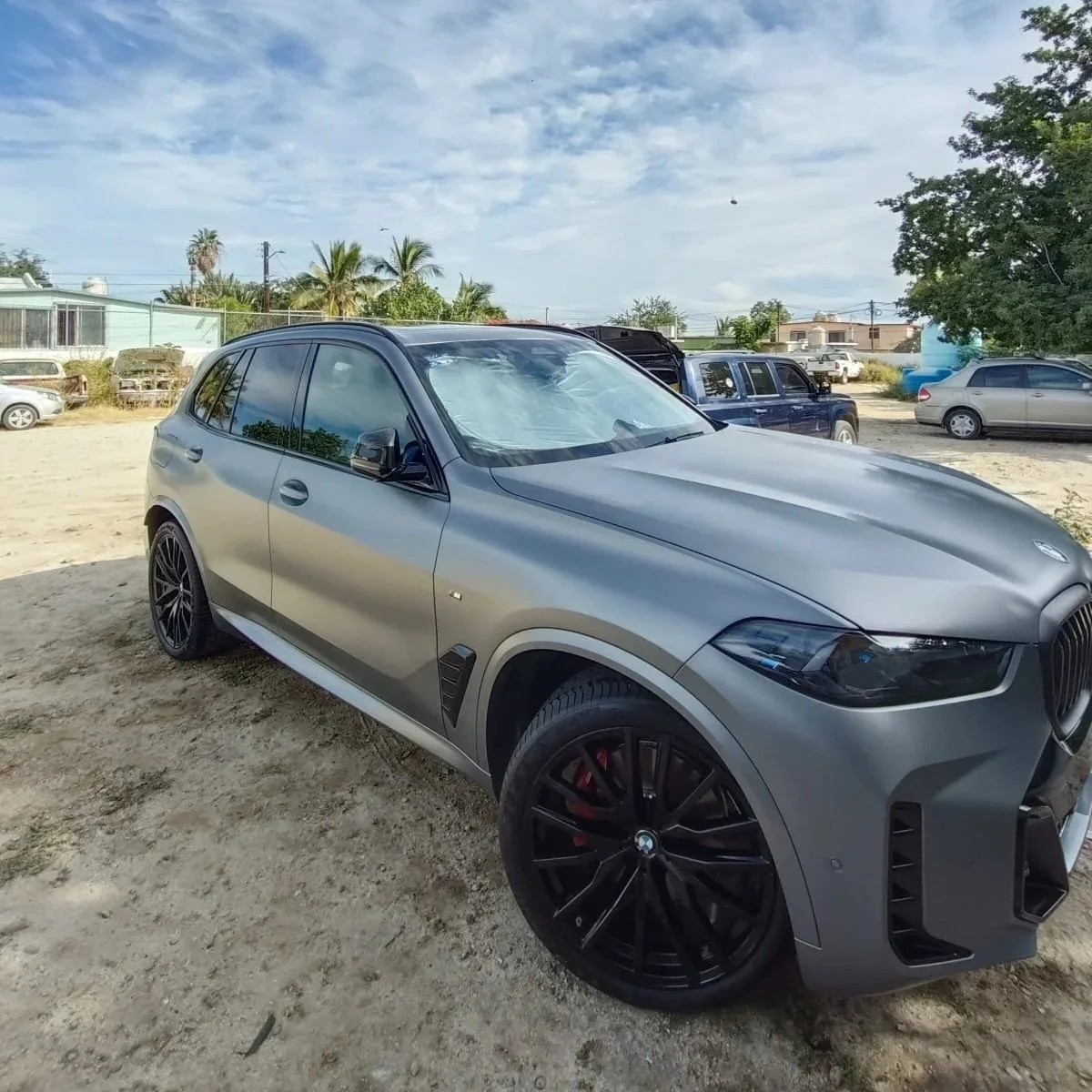 Auto gris de lujo estacionado sobre tierra en un área de lotería, con otros vehículos y árboles en el fondo bajo un cielo parcialmente nublado.