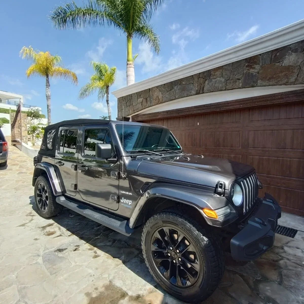 Jeep Wrangler Sahara negro con techo suave, estacionado en un patio con piedra, en un entorno con palmeras y un cielo azul con algunas nubes.