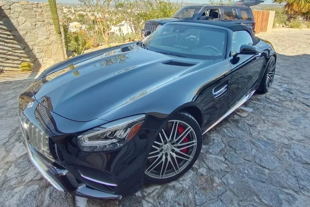 Un automóvil deportivo negro con techo rígido y llantas de diseño modernista, estacionado en un camino de piedra, con un automóvil SUV gris al fondo y vegetación en el entorno.