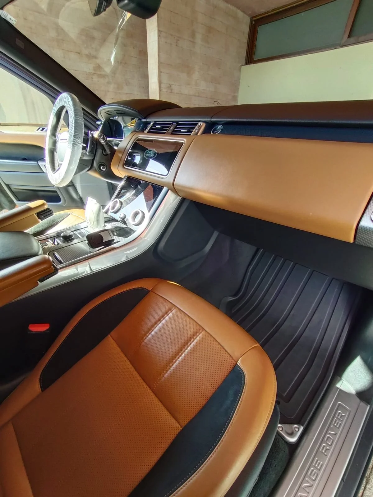 Interior de un vehículo Land Rover con asientos de cuero marrón y negro, tablero y consola central moderna, tapetes de goma negras y ventanas abiertas.