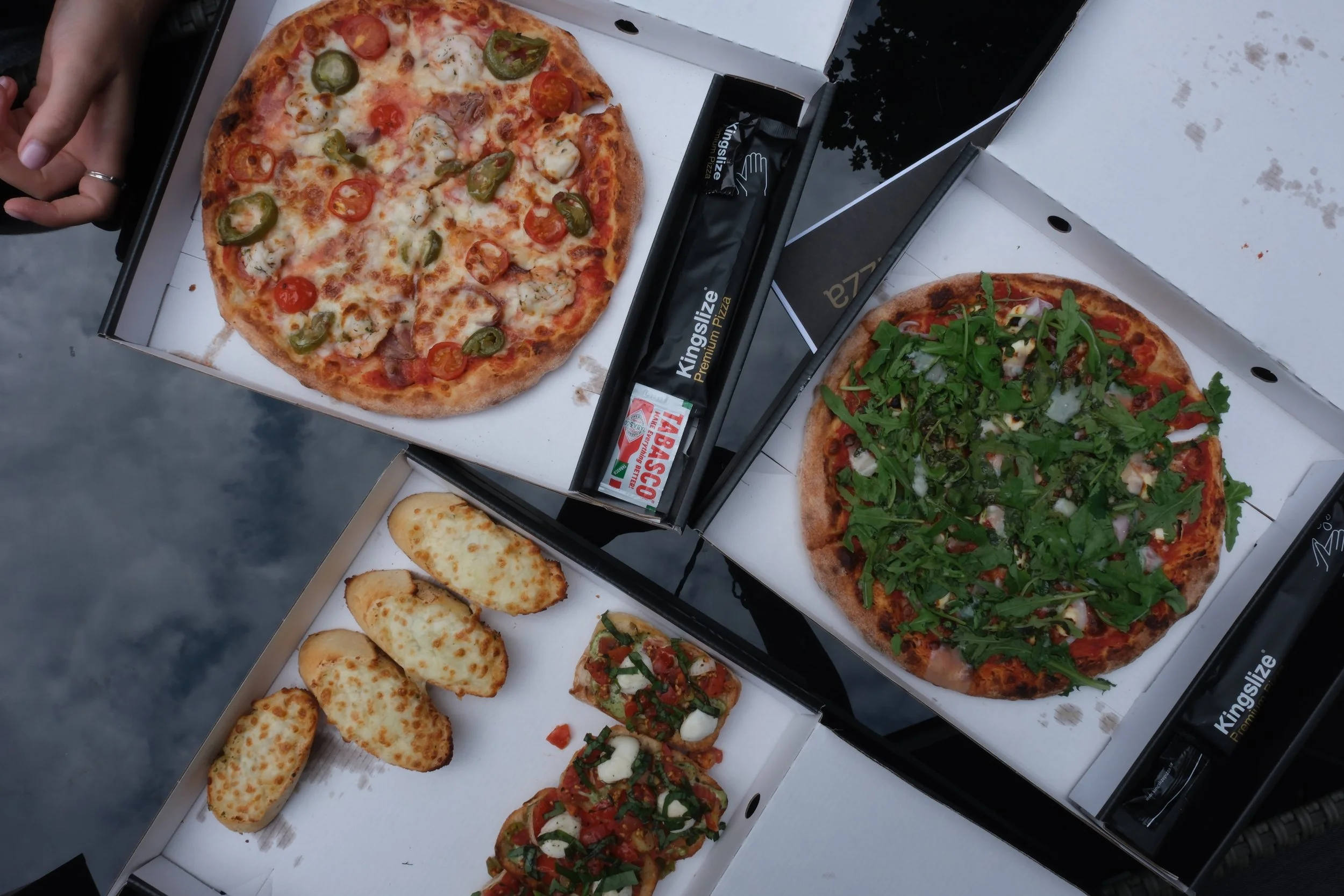 Two pizzas in boxes, one topped with jalapeños and cherry tomatoes, the other with arugula; garlic bread and a partially visible tray with various toppings. A hand is visible. Tabasco sauce packet nearby.