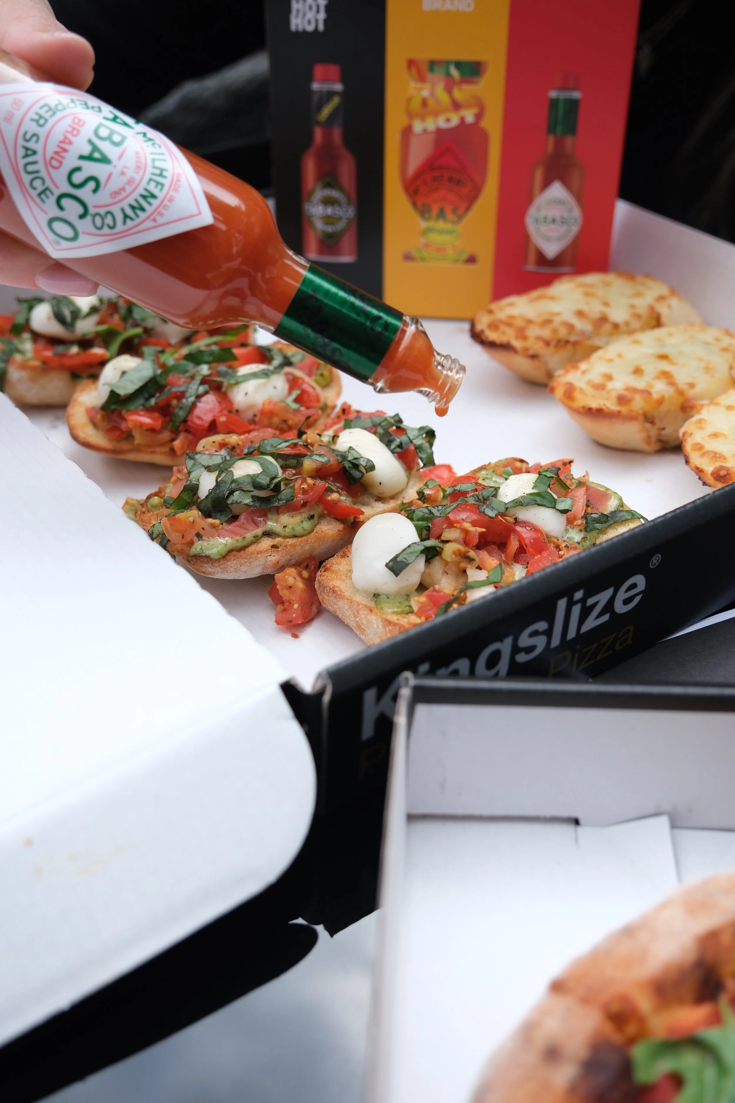 Tabasco sauce being poured onto bruschetta with mozzarella, tomato, and basil in a pizza box.