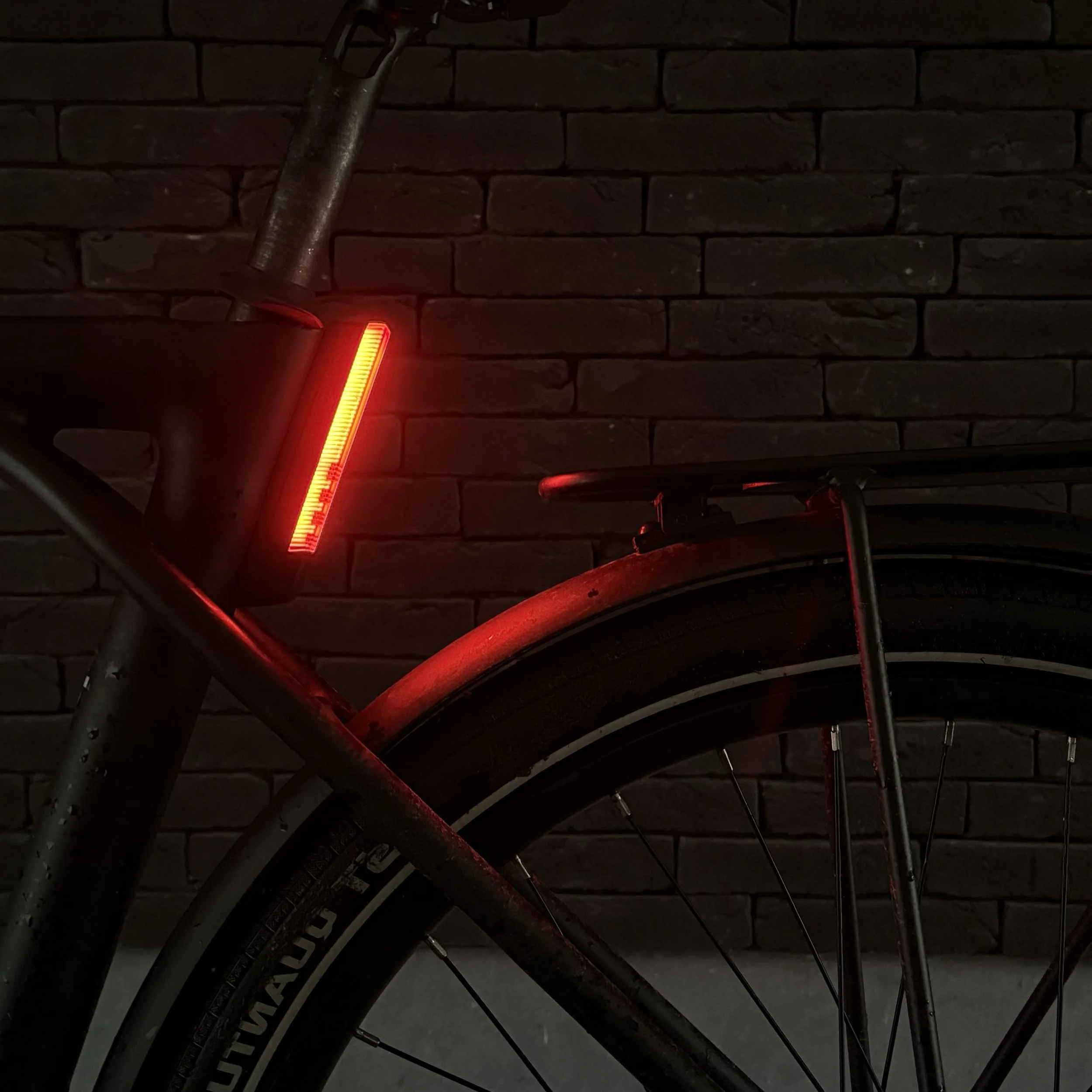 Close-up of a bicycle rear wheel and frame with a red rear light near a brick wall backdrop.