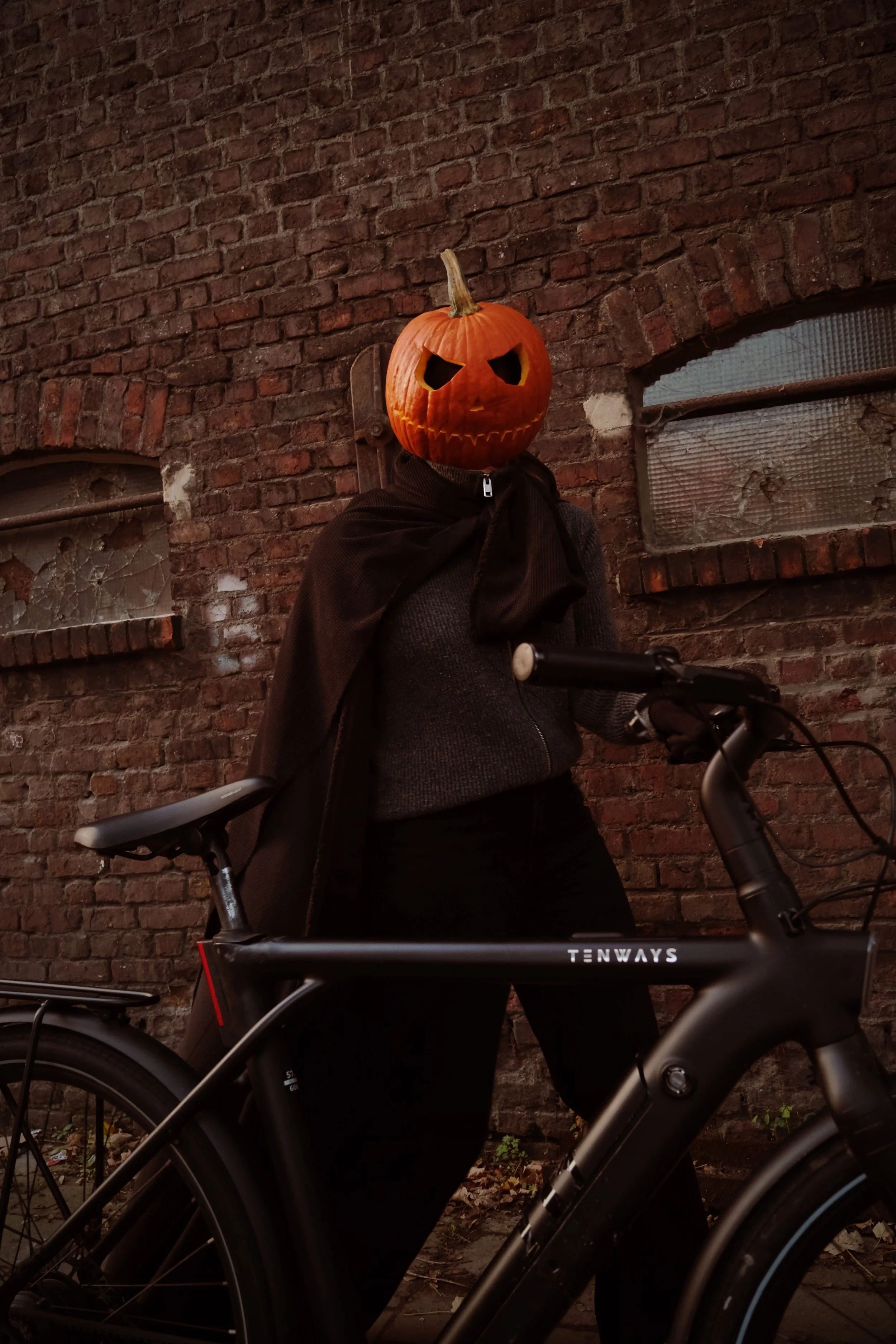 Person with a pumpkin head wearing a cape, standing with a bicycle in front of a brick wall.