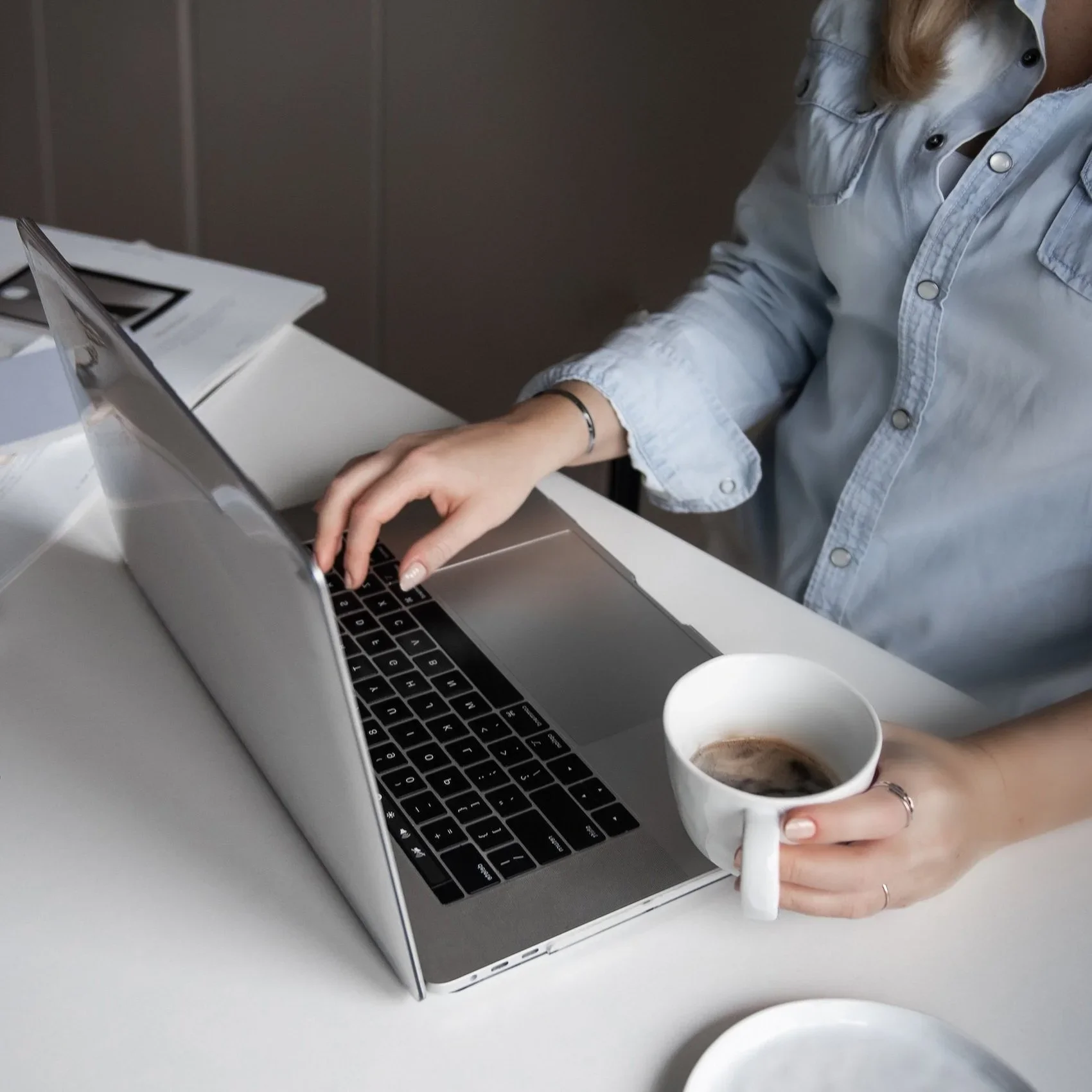 Person sitting at a desk using a laptop, holding a cup of coffee, wearing a light blue denim shirt and a bracelet.