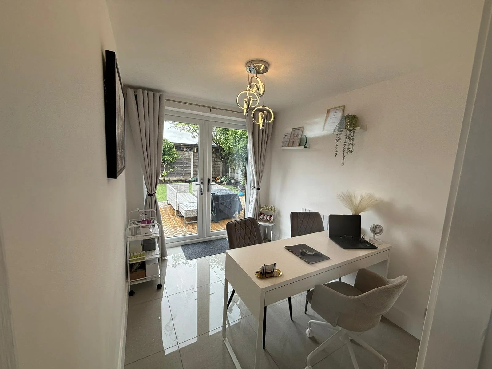 Modern dining room with white walls, a white table, and two chairs, facing glass sliding doors opening to a backyard patio with outdoor seating and greenery.