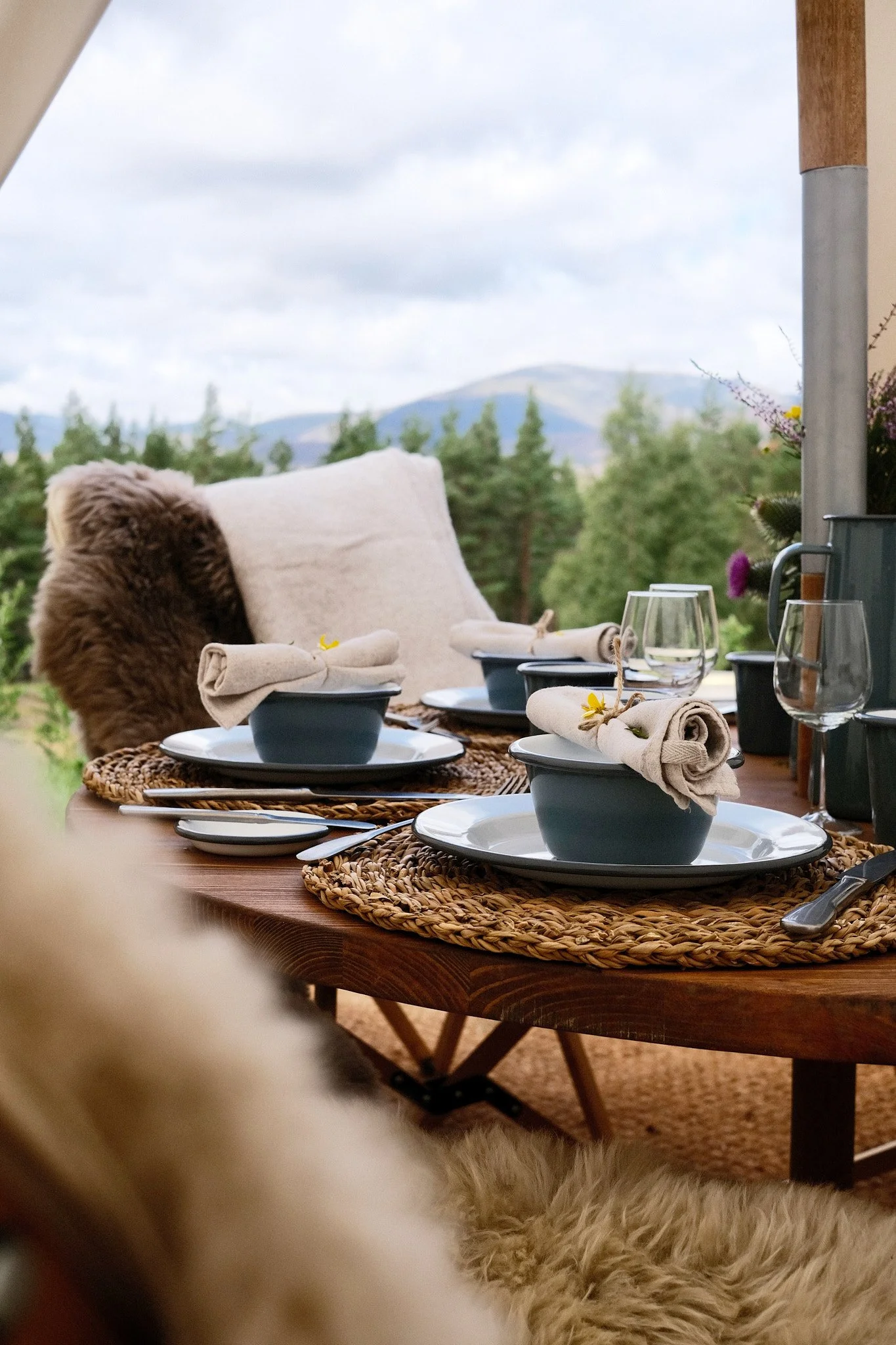 An outdoor dining table set with dishes, napkins, and glasses, surrounded by cozy cushions and placed in front of a scenic mountain view with trees and cloudy sky.