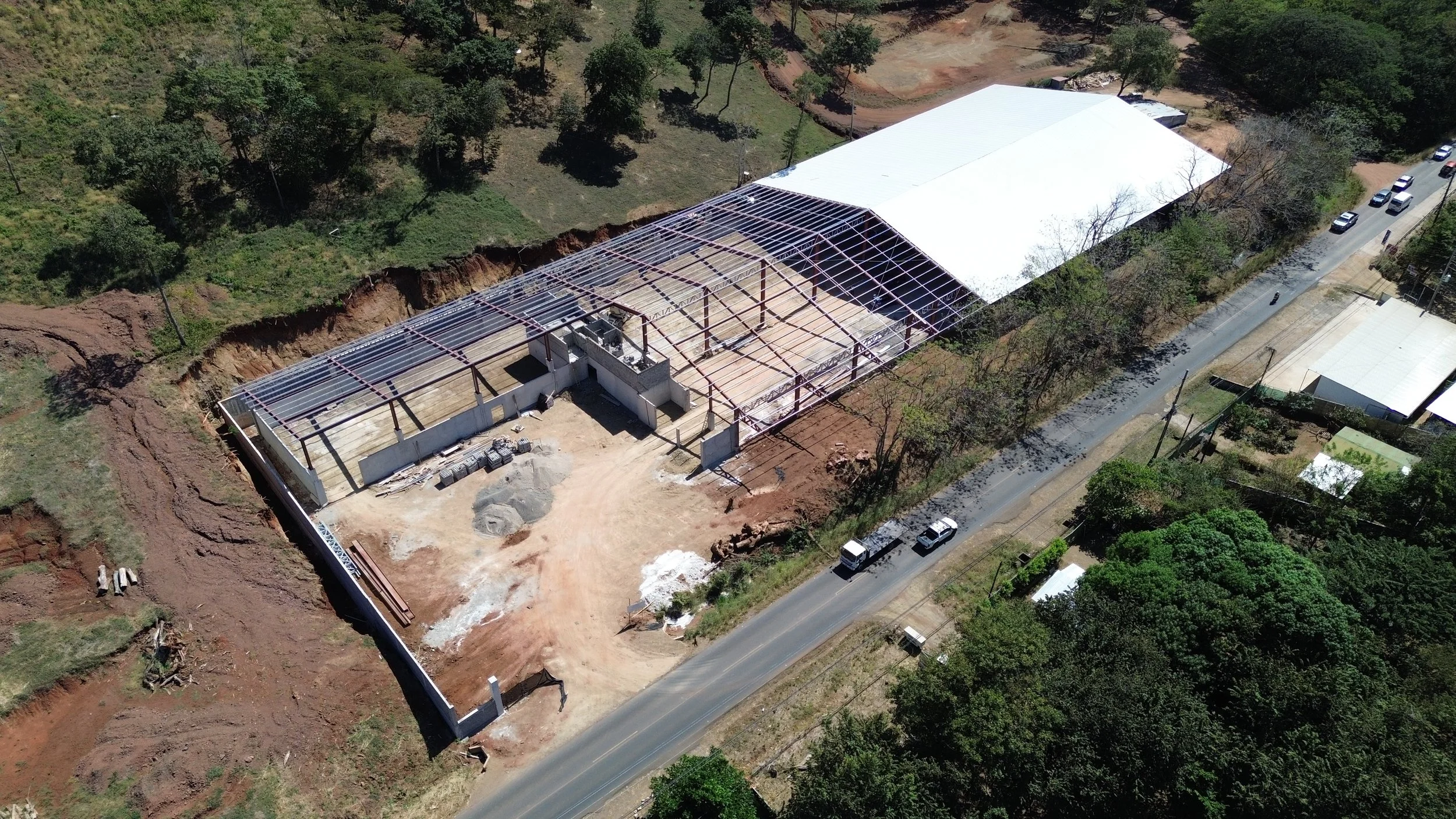 Construcción de un edificio con estructura metálica en un terreno parcialmente excavado, con un techo metálico blanco en una zona y abiertas en otra, rodeado de árboles y una calle con vehículos.