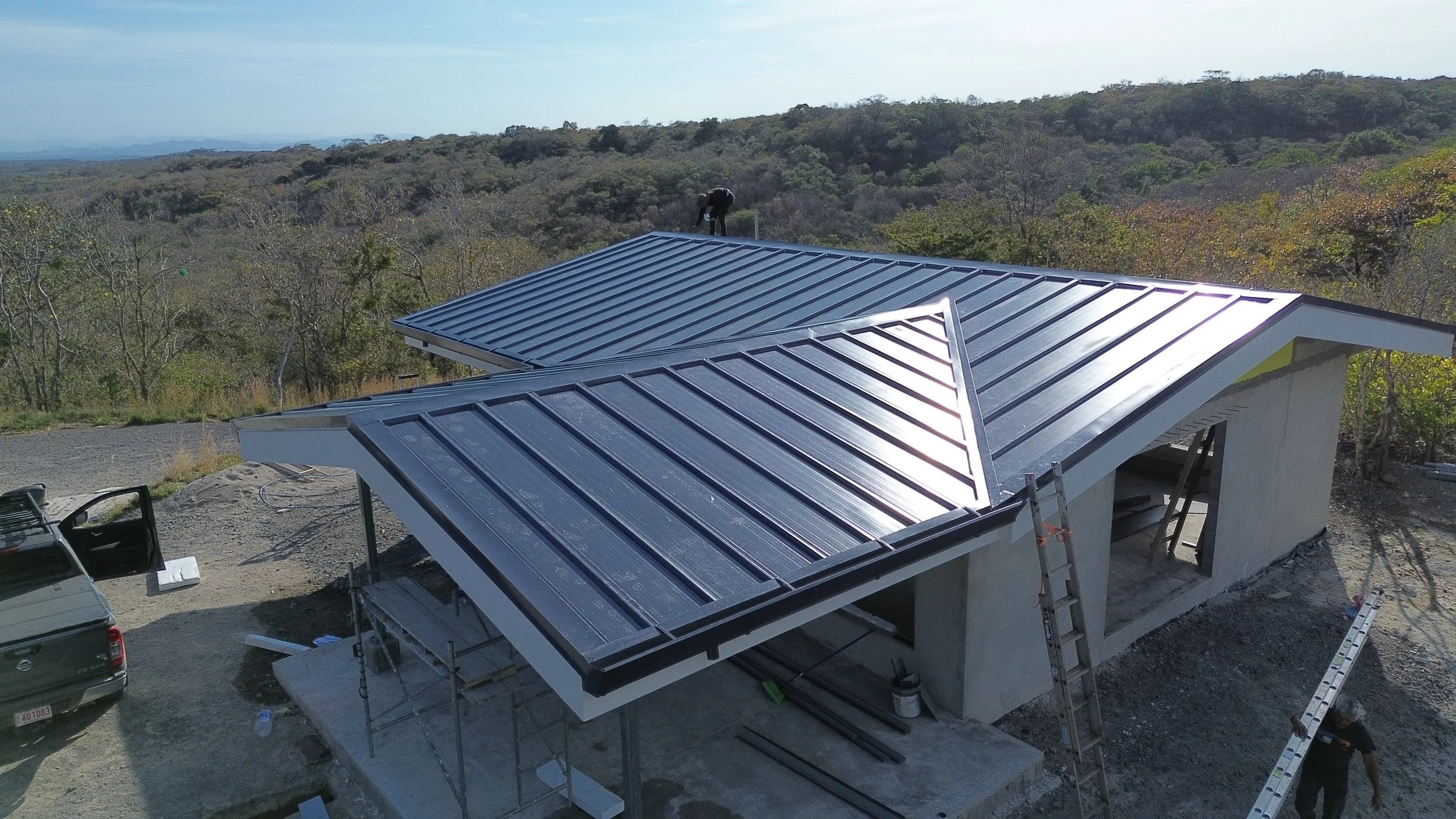 Construcción de una casa con techo de láminas metálicas en un área rural con vegetación en el fondo.