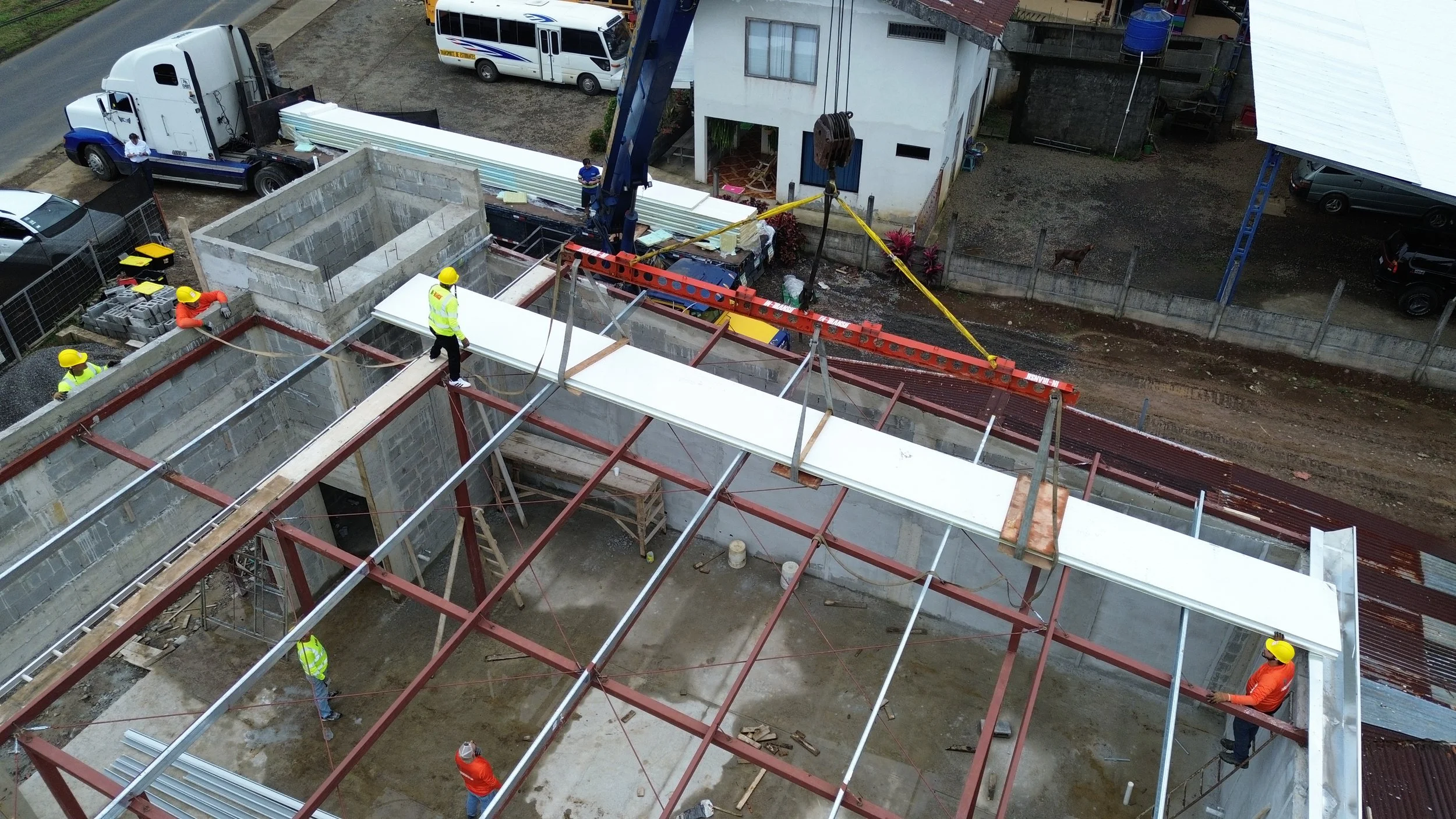 Construcción de un edificio con trabajadores en el sitio, una grúa levantando una losa y varios vehículos y casas cercanas.