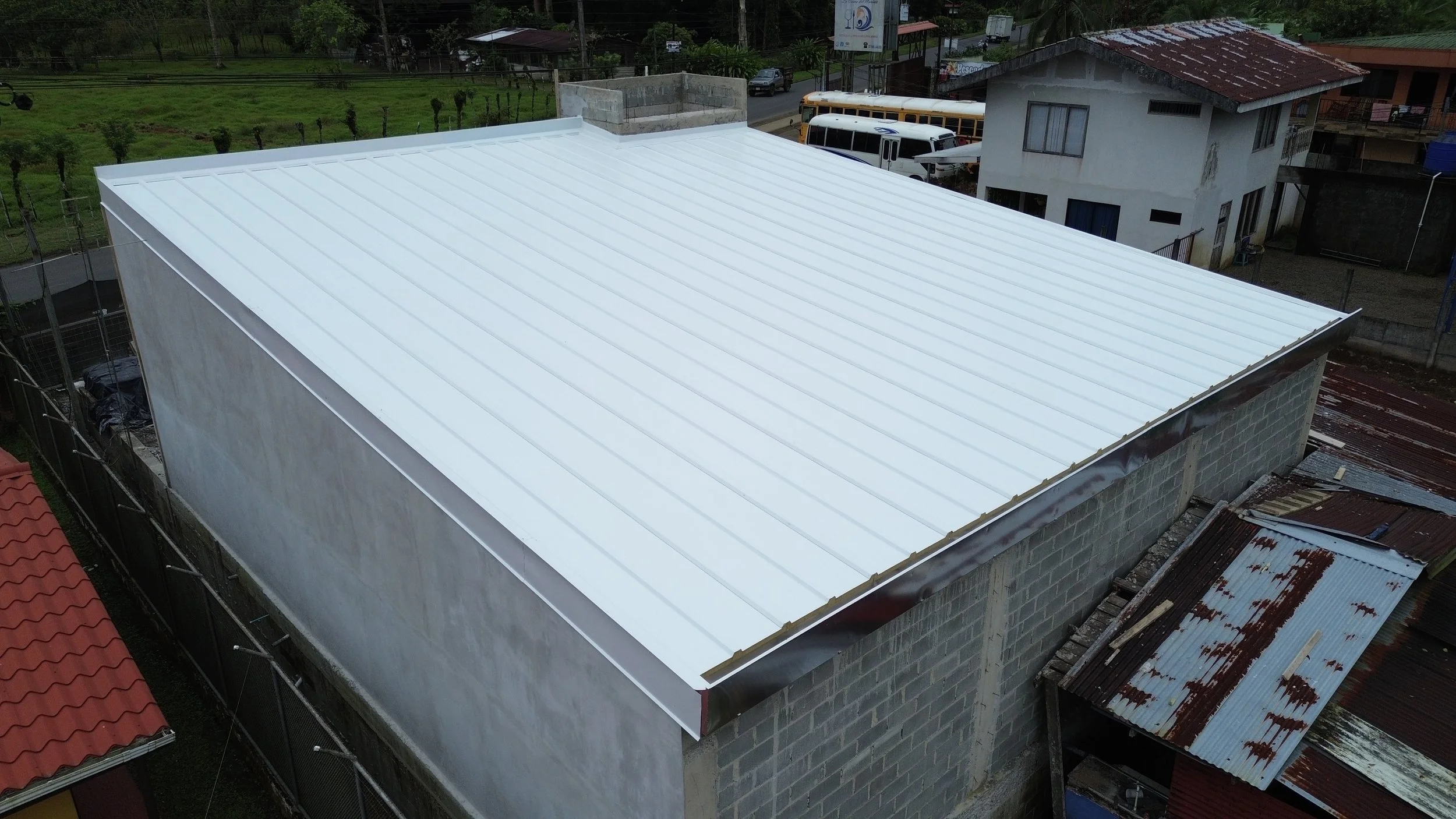 Vista aérea de un techo de lámina metálica blanca en construcción sobre un edificio, con casas y árboles en el fondo.