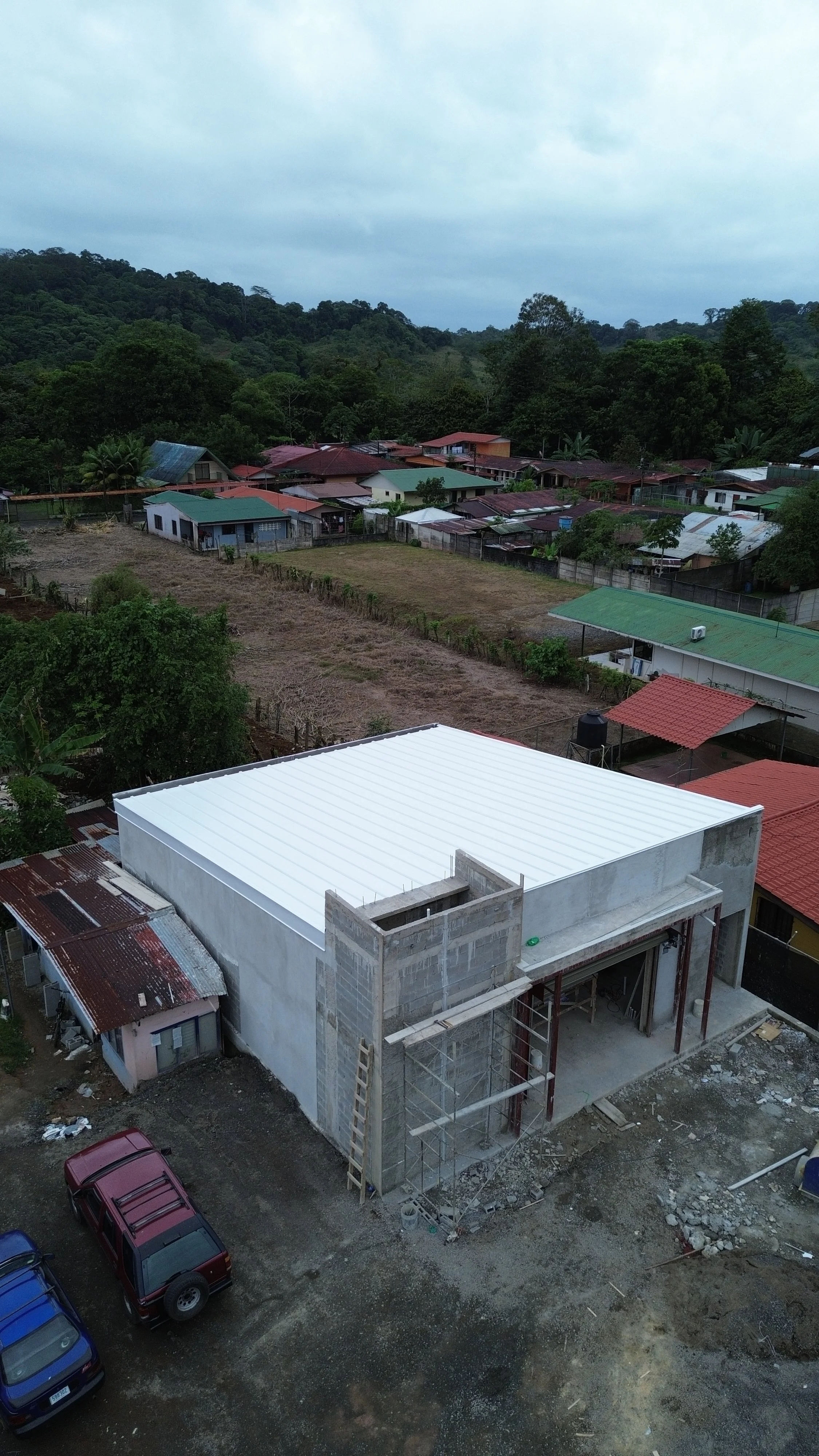 Construcción de un edificio en un área rural, con paredes de bloque y techo de lámina blanca, vehículos estacionados y casas dispersas en un entorno boscoso.