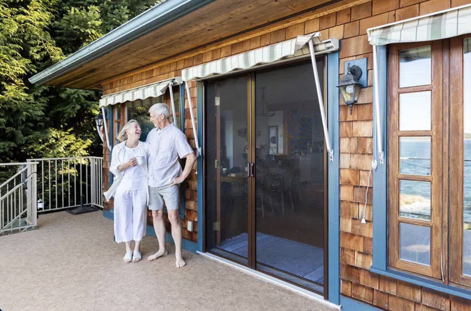 Retractable screen door framing an elderly couple smiling on a balcony beside a wooden house with large windows and awnings.