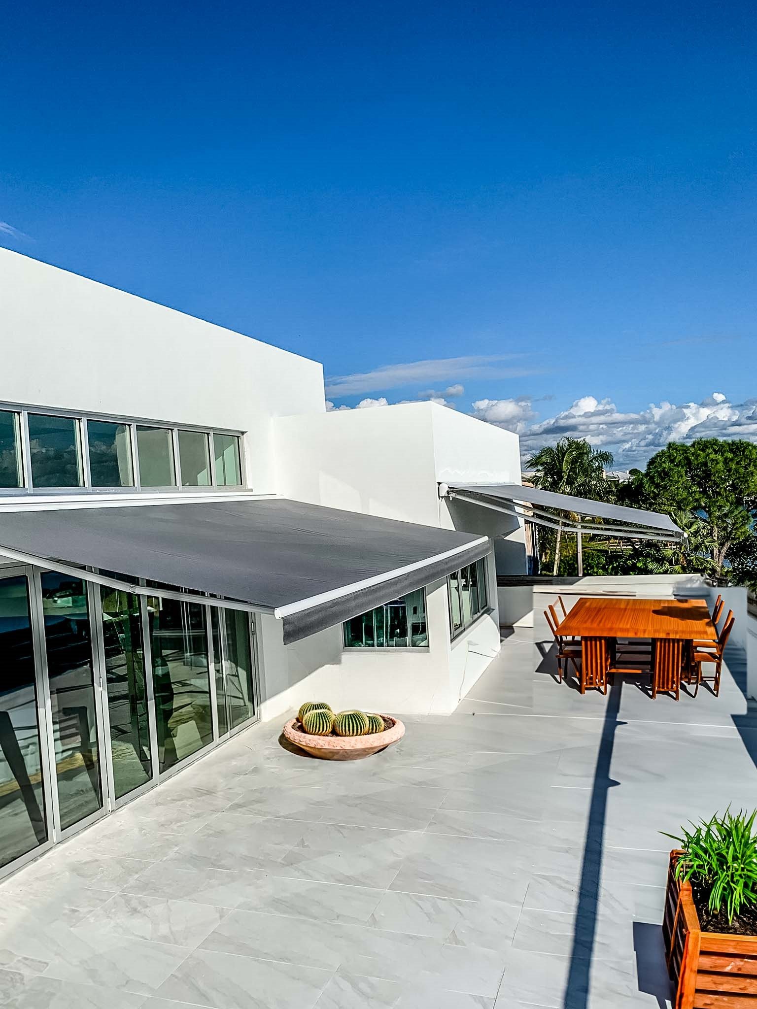 Modern patio with retractable awning, glass doors, wooden dining table and chairs, potted cacti, and clear blue sky.