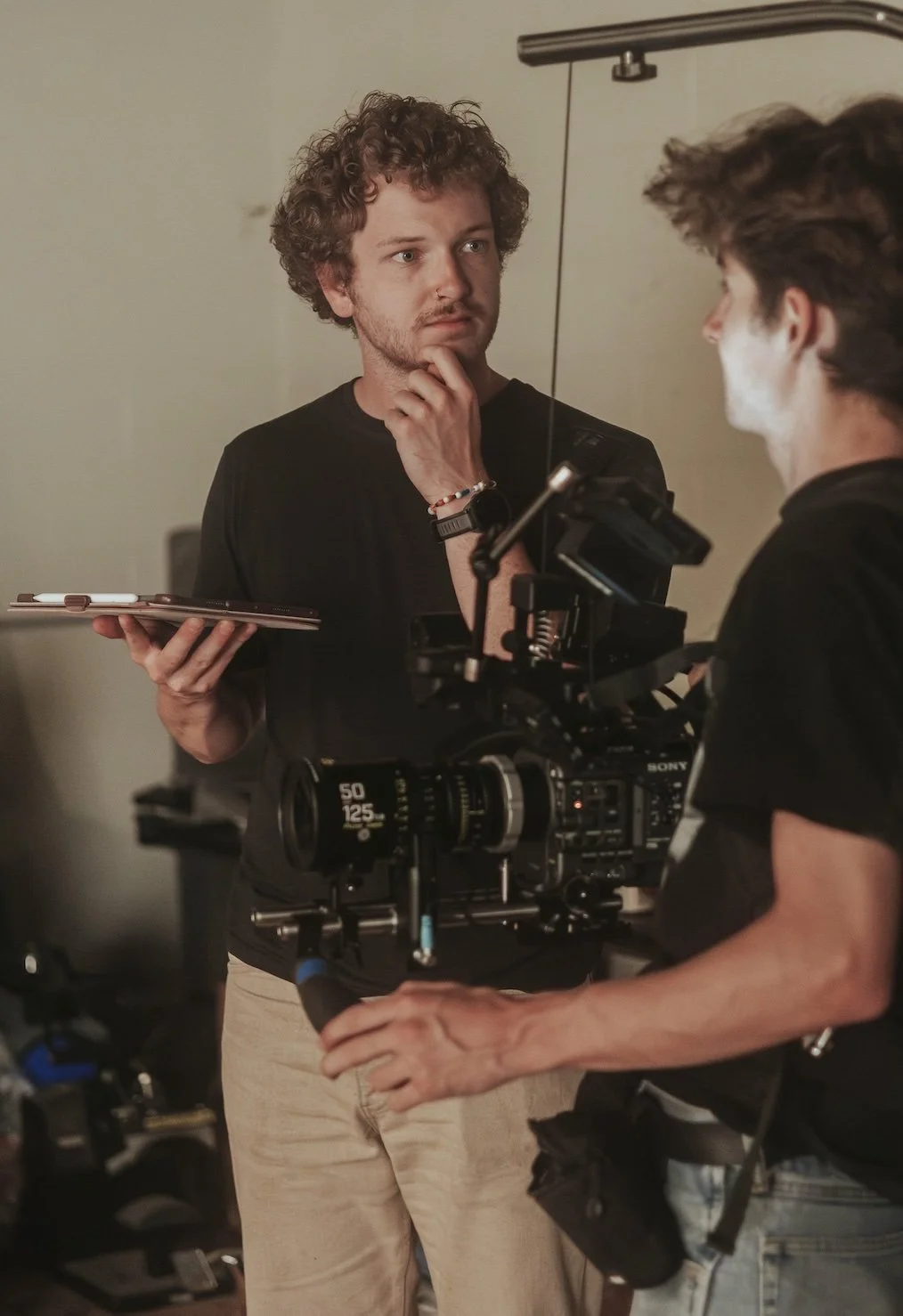 Two men on a film set, one holding a clipboard and the other operating a professional camera. The man operating the camera has makeup on his face.