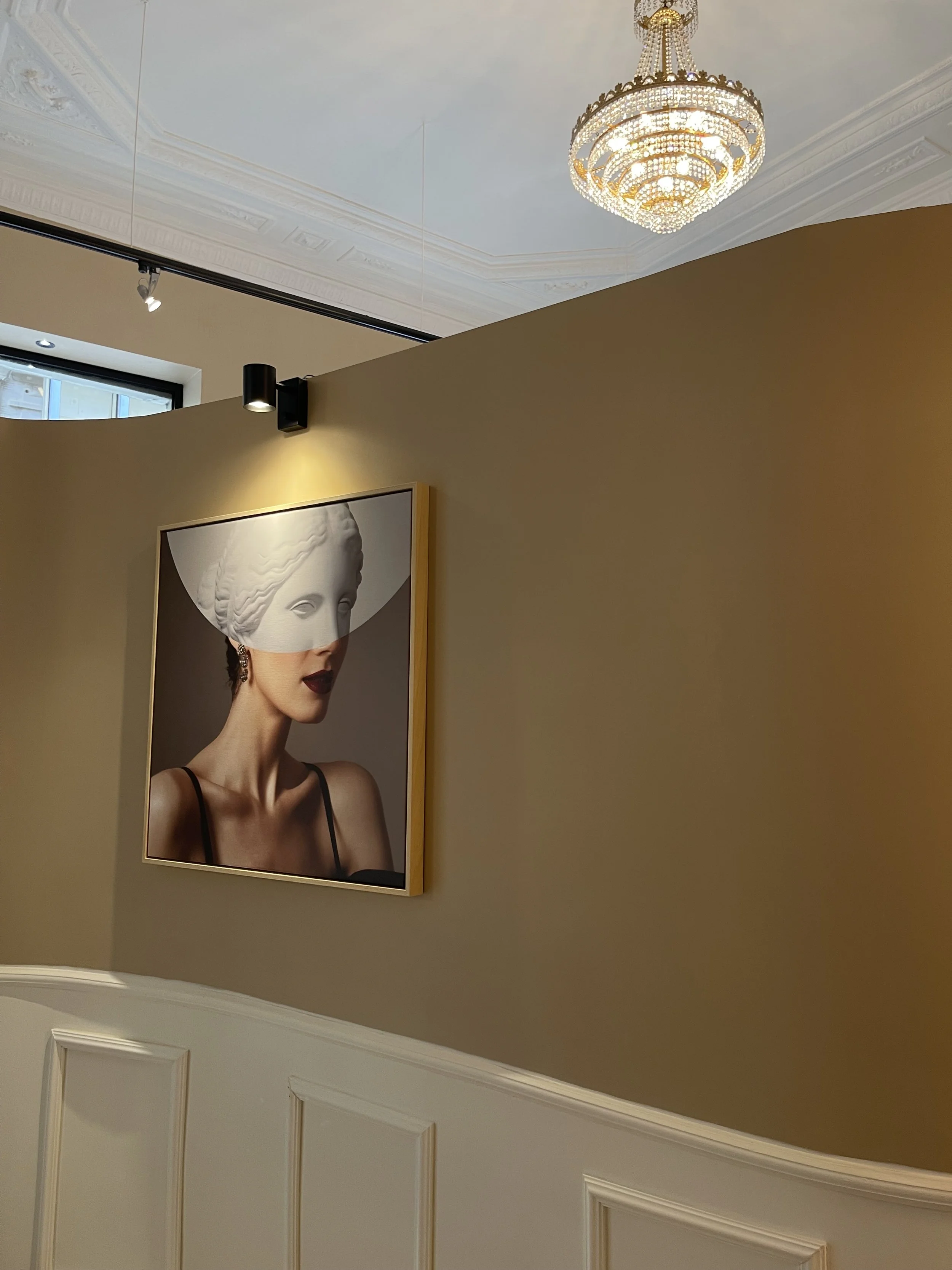 Une photo d'art moderne montrant une femme avec un visage partiellement caché par une sculpture classique. L'image est accrochée sur un mur beige, sous un lustre élégant, dans une pièce au plafond décoré.
