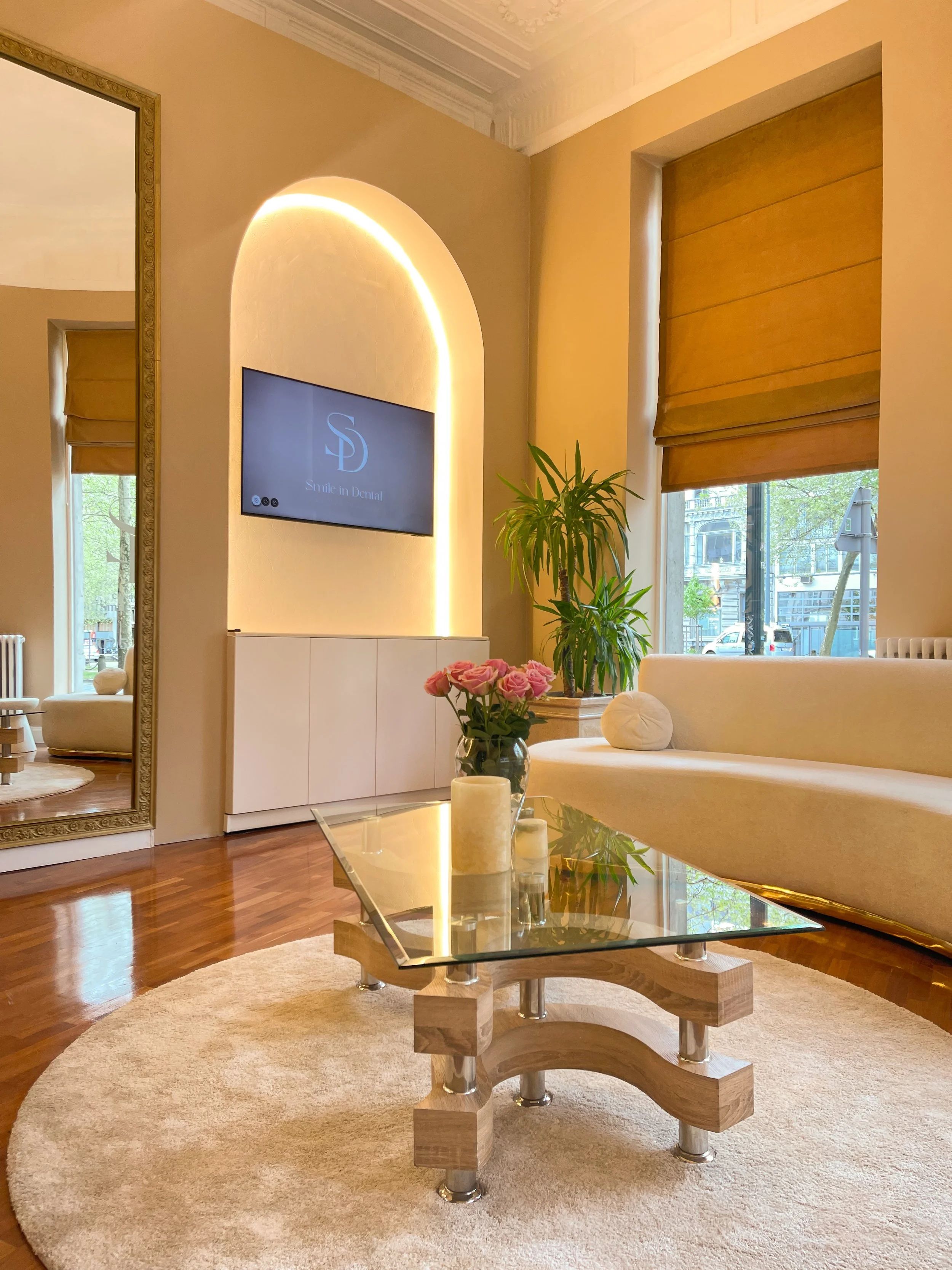 Salon moderne et lumineux avec un canapé courbé beige, une table basse en verre, un vase de roses sur la table, et une télévision accrochée au mur. Il y a des plantes vertes et un grand miroir. Les murs sont beige clair et une fenêtre avec store roma