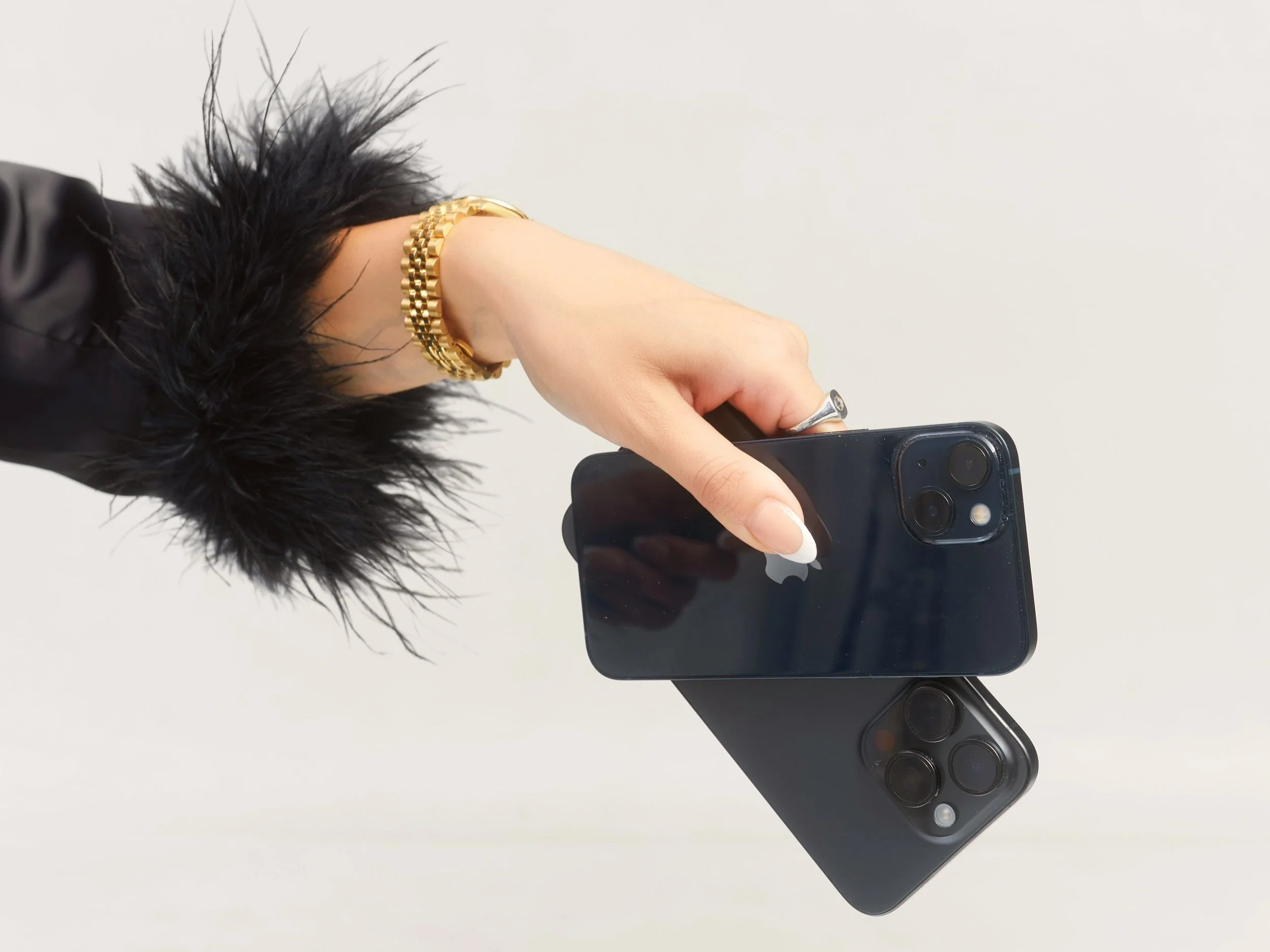 A person holding two black iPhones with one hand, wearing a gold bracelet and a silver ring, against a plain light background.