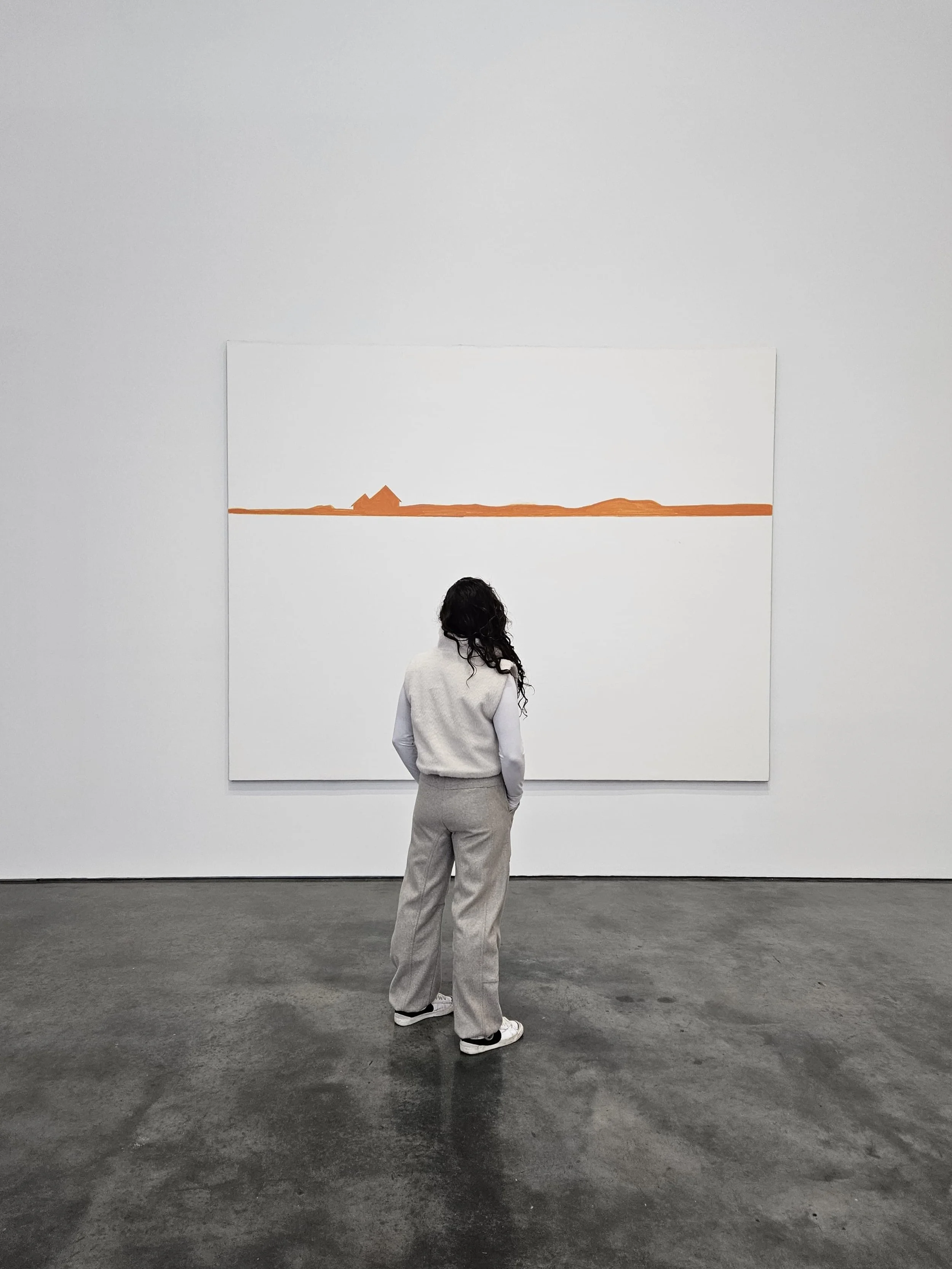 A woman stands in an art gallery viewing a large white painting with orange lines and shapes resembling a horizon and landforms.