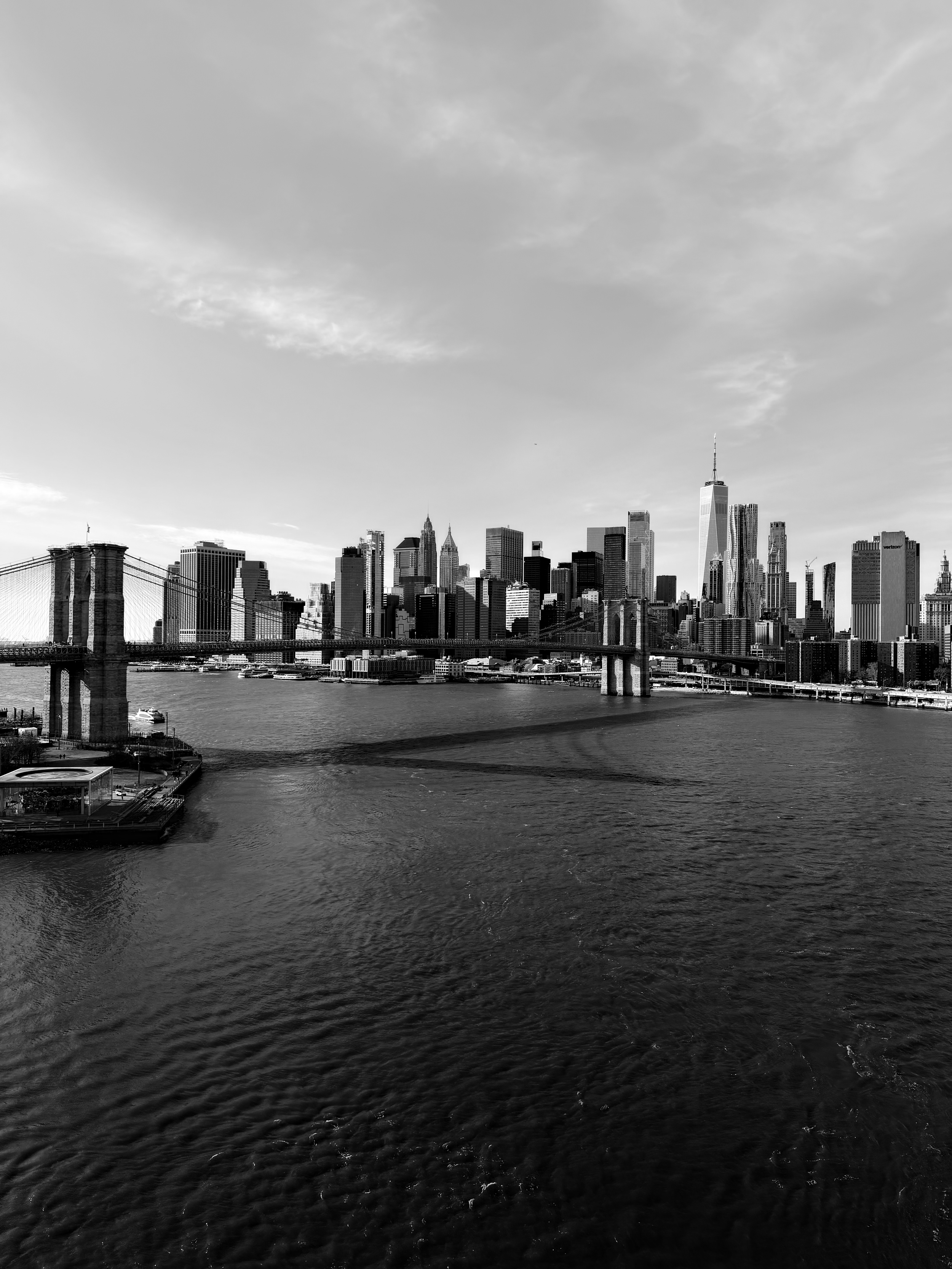 Black and white photo of the New York City skyline, including the Brooklyn Bridge over the East River with boats and water in the foreground.