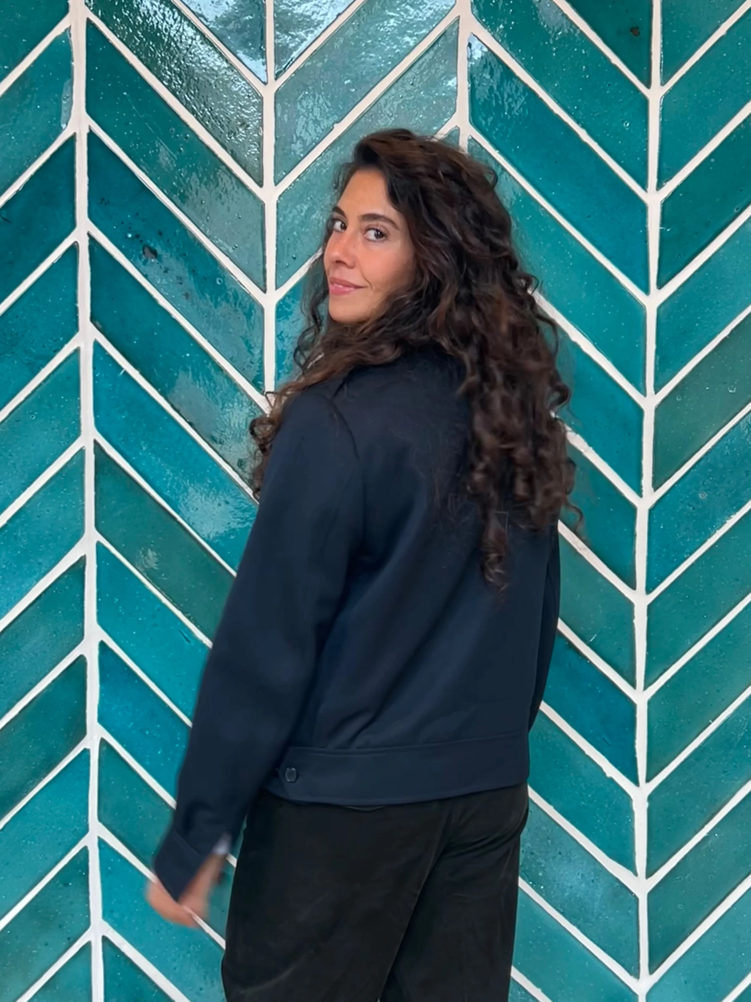 A woman with long curly brown hair, wearing a dark jacket and black pants, standing against a teal wall with white geometric patterns