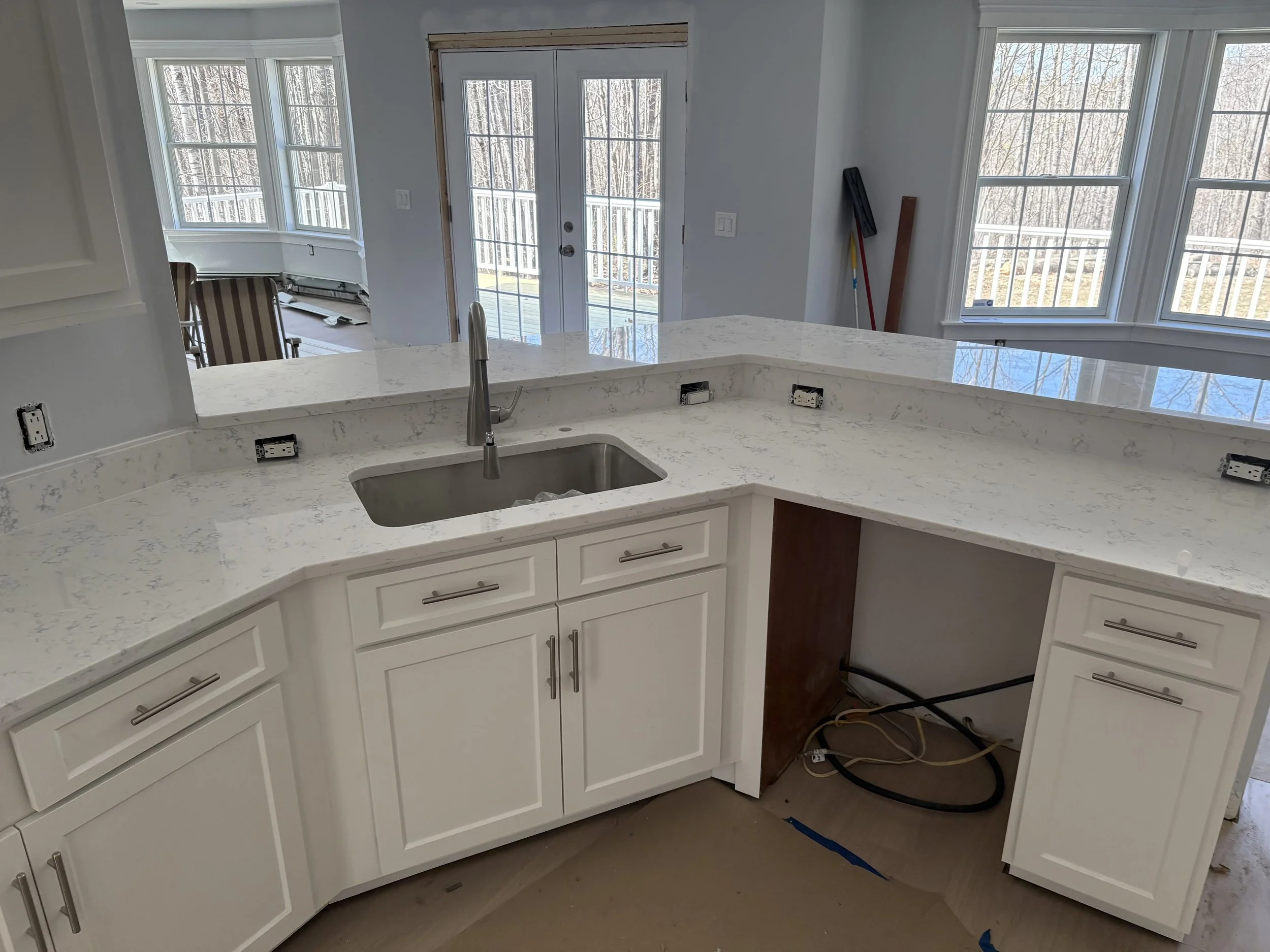 Kitchen with white cabinets, a marble countertop, and a stainless steel sink. There are unfinished electrical outlets and some construction tools and materials nearby. Large windows and a door leading to an outdoor deck are visible in the background.