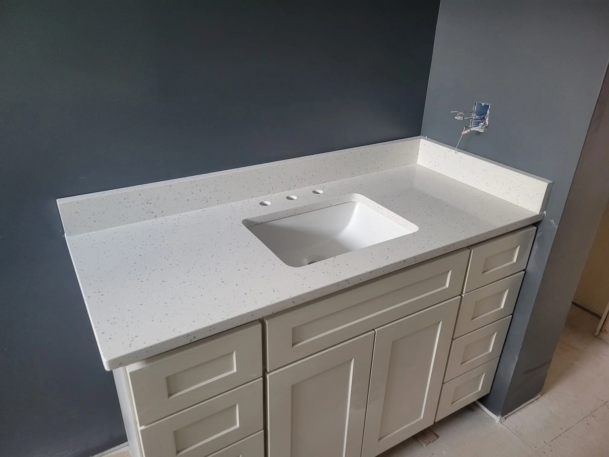 Bathroom vanity with a white speckled countertop, built-in sink, and white cabinet drawers below, with a dark gray wall behind.