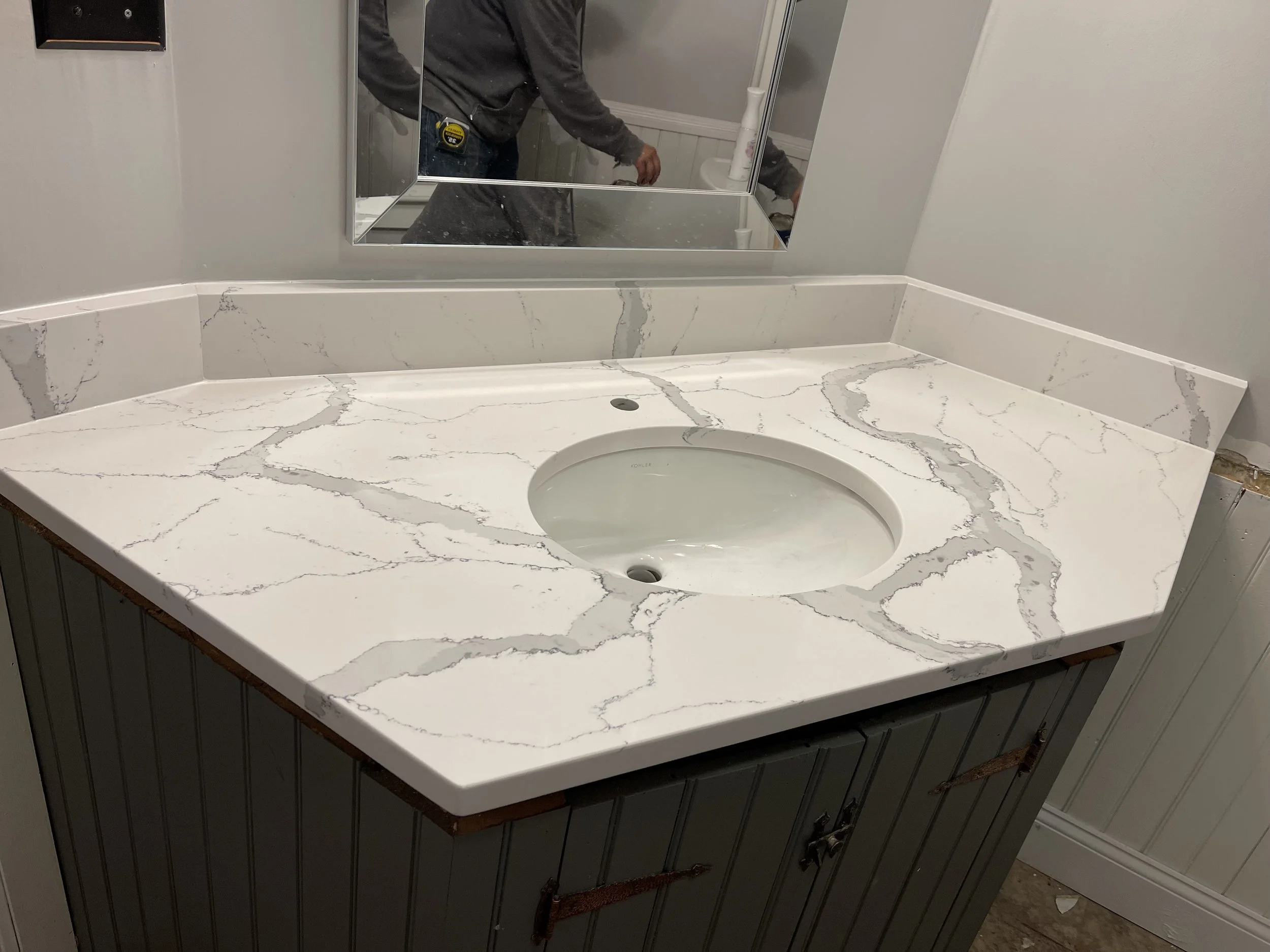 A bathroom countertop made of white marble with gray veining, featuring an open sink and a bathroom mirror reflection showing a person in a gray hoodie holding a phone or camera.