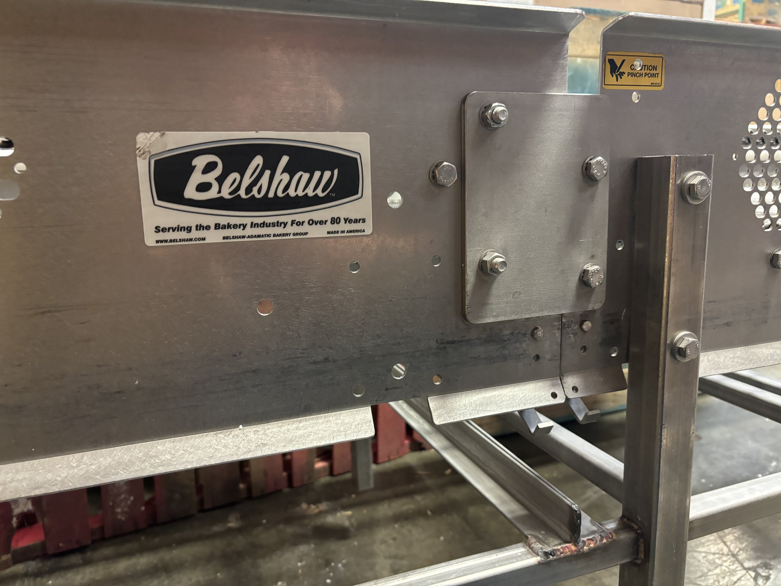 Close-up of industrial metal machinery with "Belshaw" brand label and caution sticker, featuring bolts and metal framework.