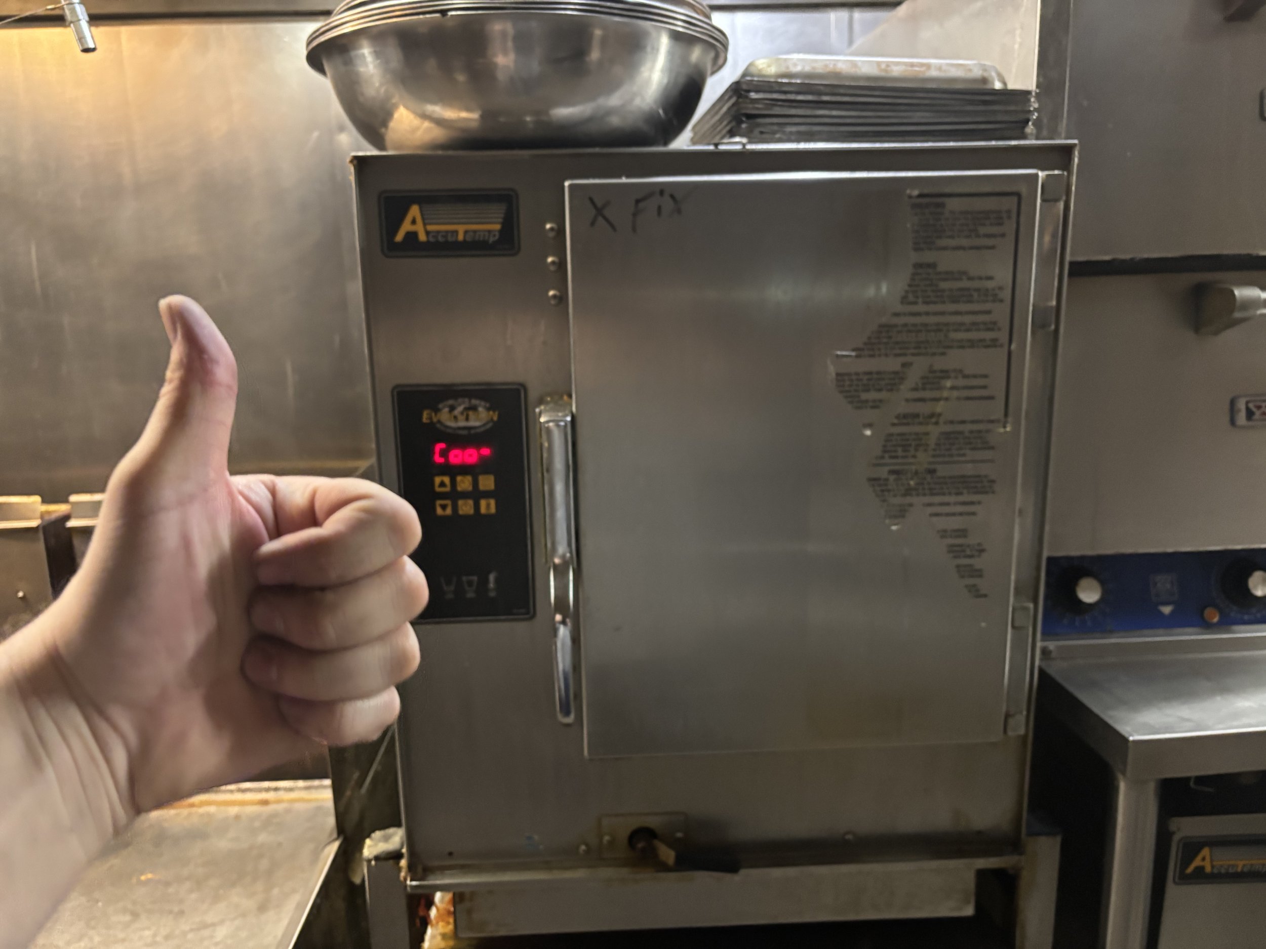 Hand giving a thumbs-up in front of a commercial kitchen steamer with digital controls and metal surface on top.
