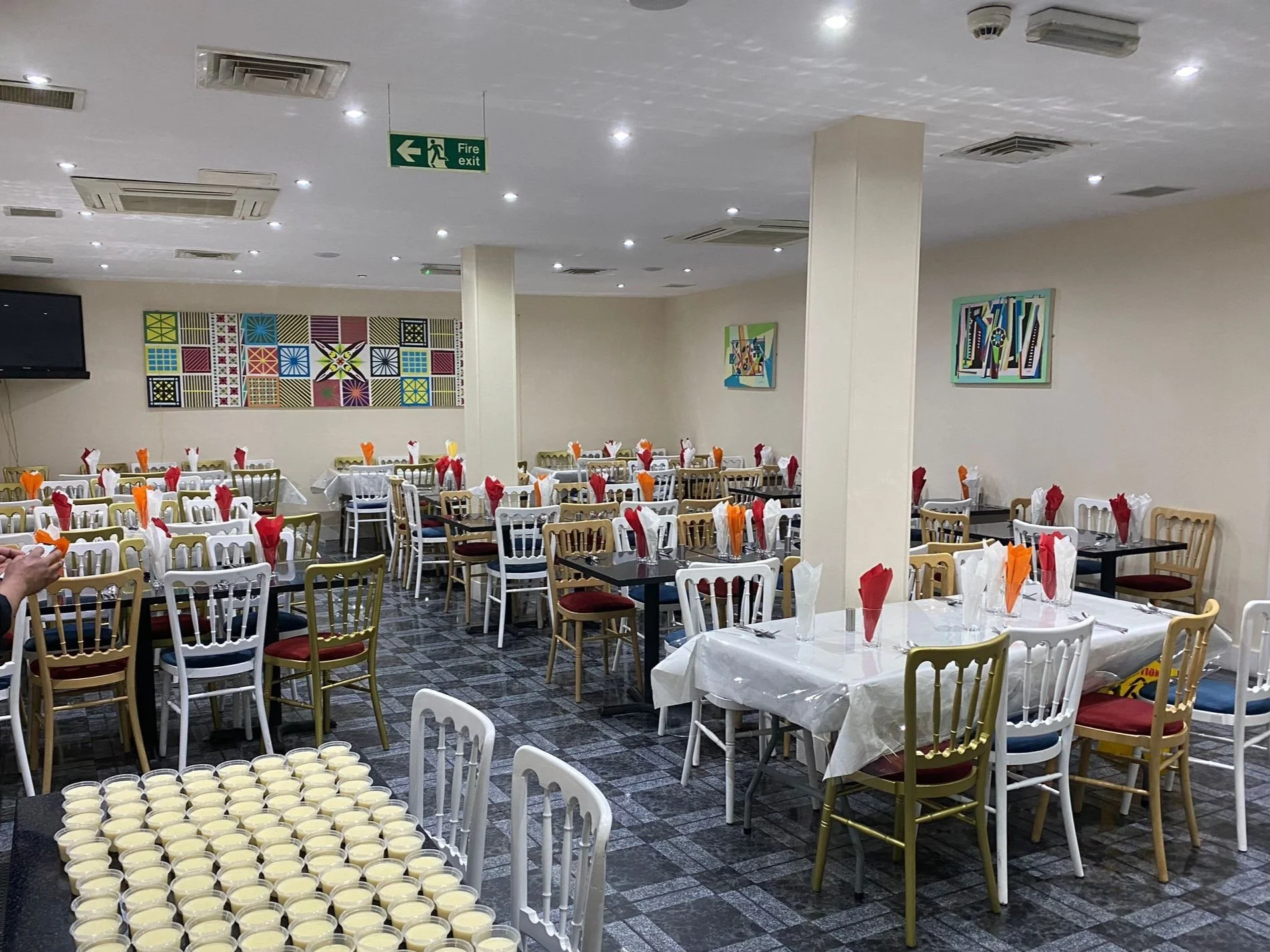 Interior of a large dining area with multiple tables set with colorful napkins and white tablecloths. The chairs around the tables are gold and white with red or blue seats. A stack of small dessert cups is in the foreground. The walls display modern artwork and there is a 'fire exit' sign above a door.