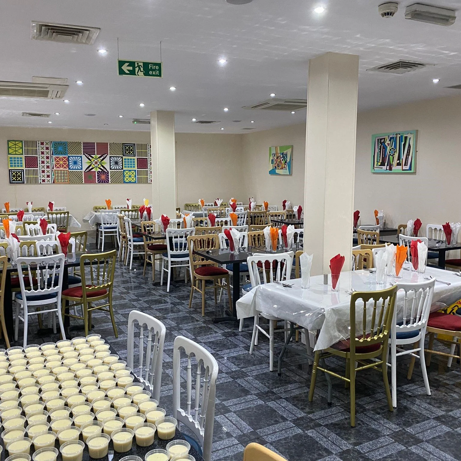 A spacious dining room set up for an event, featuring multiple tables with arranged chairs adorned with red, white, and orange napkins. The room has patterned tile flooring, colorful wall art, and numerous glasses filled with a creamy beverage neatly
