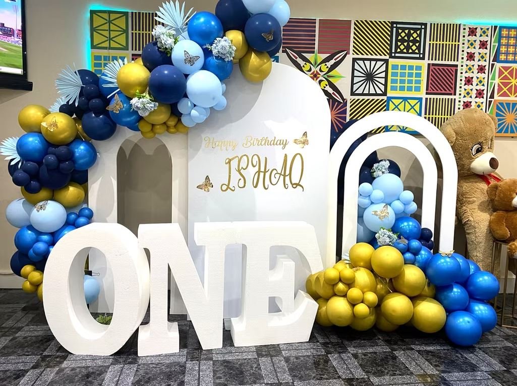 Birthday party setup with large letters spelling 'ONE', surrounded by blue and gold balloons, and a decorative backdrop with colorful patterns. A teddy bear is visible on the right.