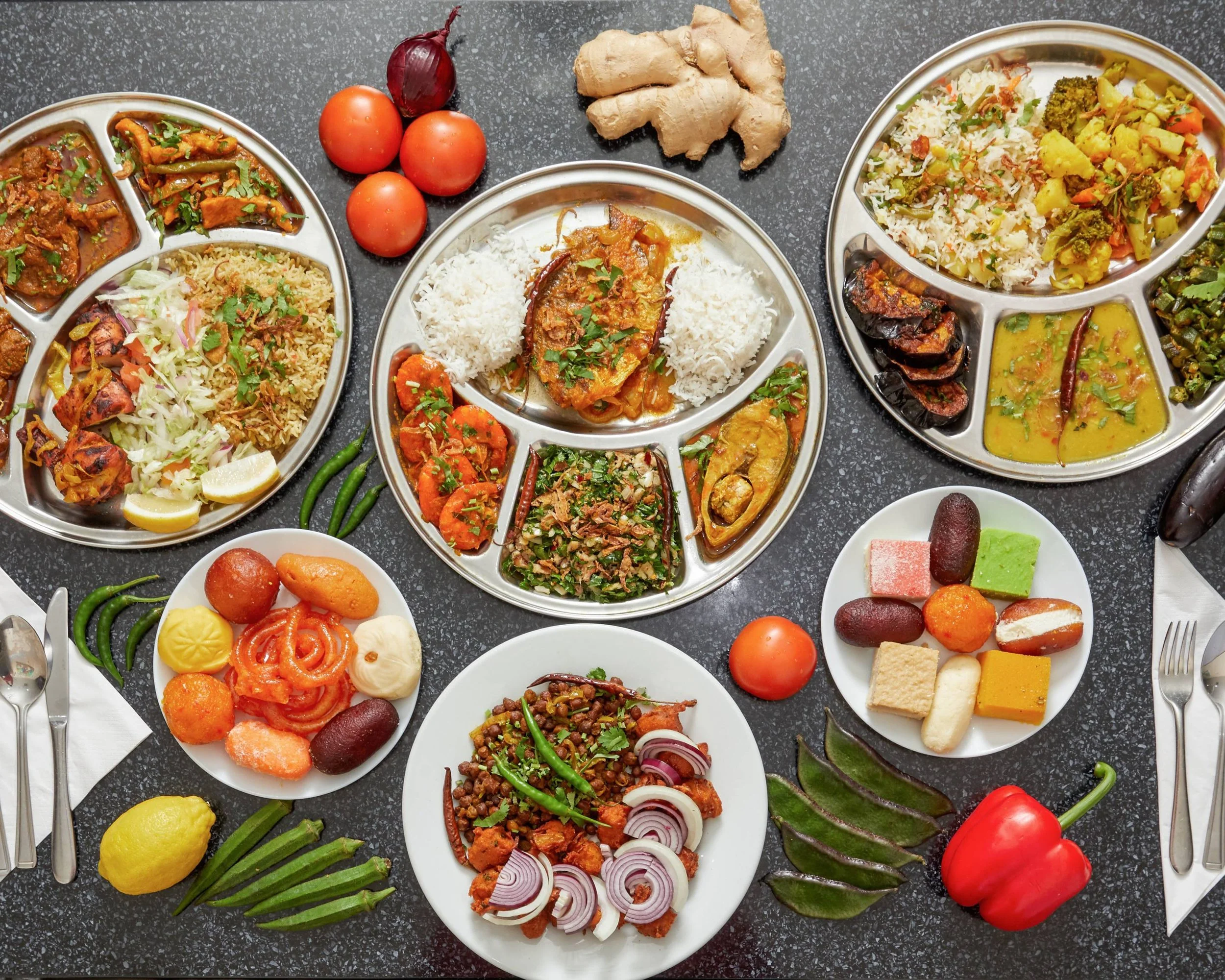 Assorted Indian dishes on a table, including thalis with rice, curry, dal, vegetables, naan, and a variety of colorful Indian sweets. Fresh vegetables like tomatoes, ginger, bell peppers, and green chilies are also present.