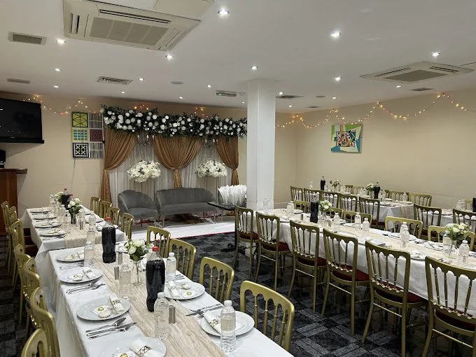 Banquet hall with white tablecloths, set tables, golden chairs, floral decorations, and string lights on walls.