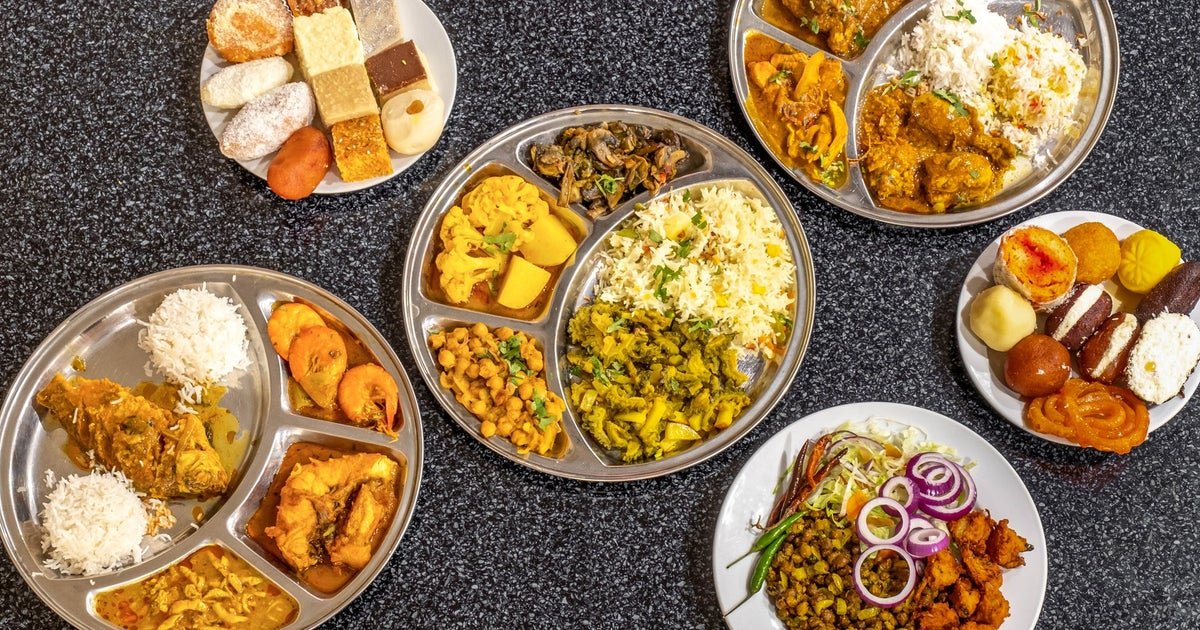 An assortment of Indian thali meals and desserts on a dark countertop. Several metal plates with compartments containing rice, curries, vegetables, and fried items. Two white plates display a variety of Indian sweets and snacks.