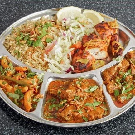A stainless steel platter with various Indian dishes including biryani rice, tandoori chicken, vegetable curry, and two types of meat curries garnished with cilantro, accompanied by salad and lemon slices.