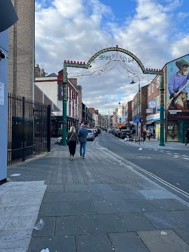 Street view with arch in the distance, people walking, shops lining the road, and a mural on the brick wall. Cloudy sky overhead.