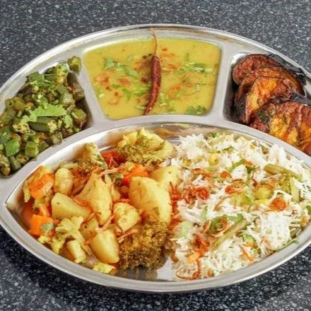 Traditional Indian thali with rice, mixed vegetable curry, okra, dal, and fried eggplant slices on a metal plate.