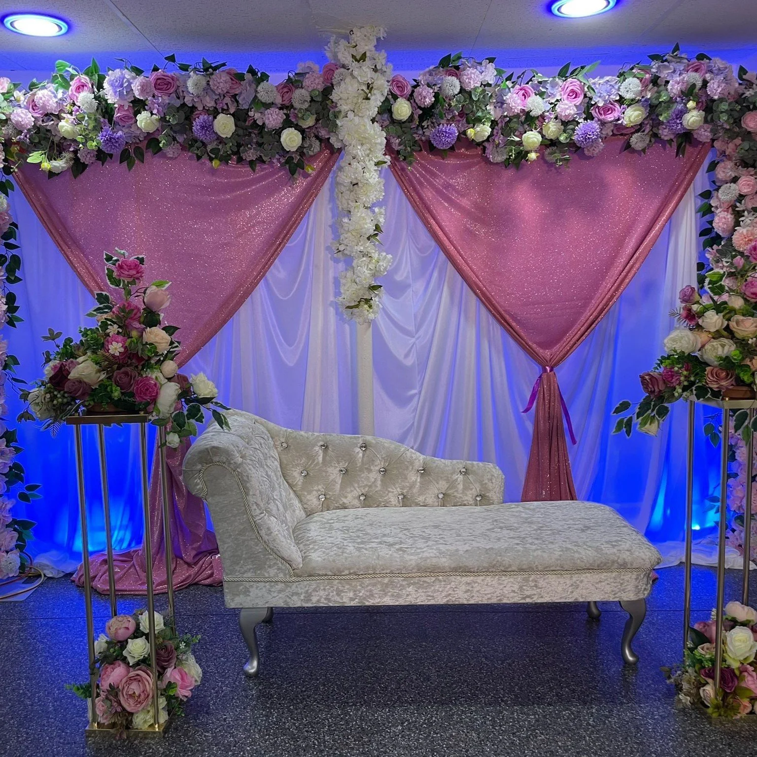 Decorative setting with a floral backdrop featuring pink drapes, white flowers, and a white tufted chaise lounge. Floral arrangements with pink and white roses are on tall stands.