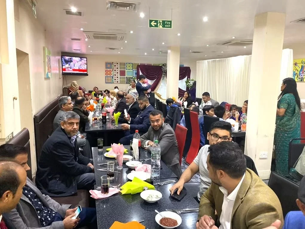 A group of people gathered inside a restaurant, seated around a long table with food and drinks. The setting is casual with bright lighting and a TV on the wall displaying a program. The atmosphere appears festive and there are decorations and flower