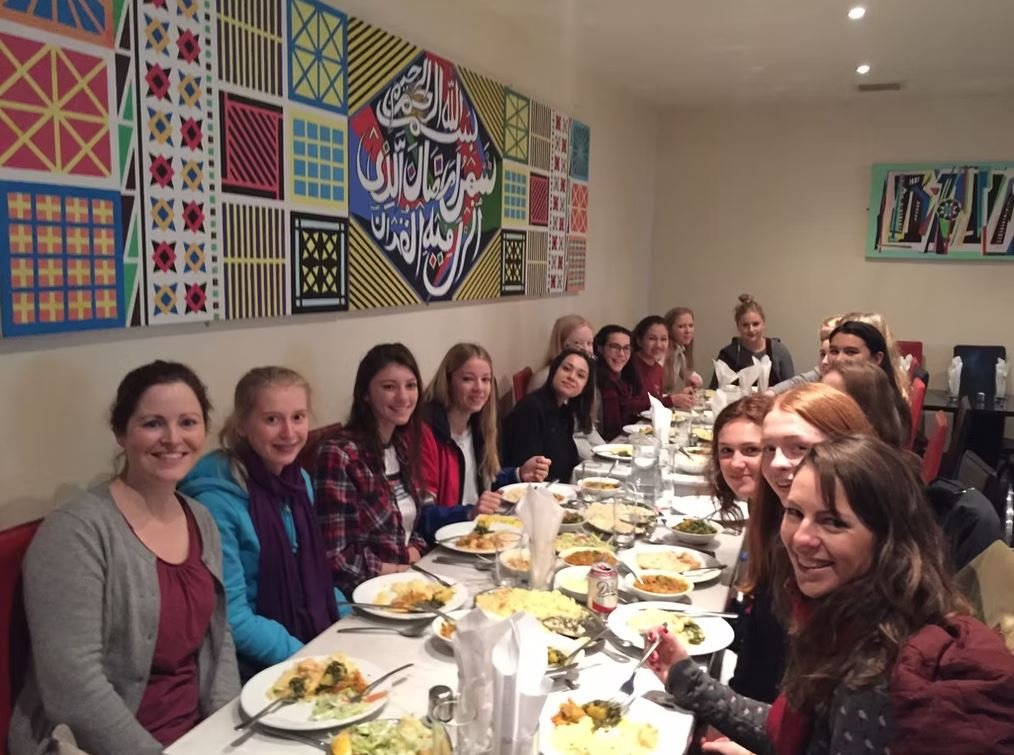 Group of people sitting at a long table in a restaurant, enjoying a meal together. The wall features colorful artistic patterns and Arabic calligraphy.