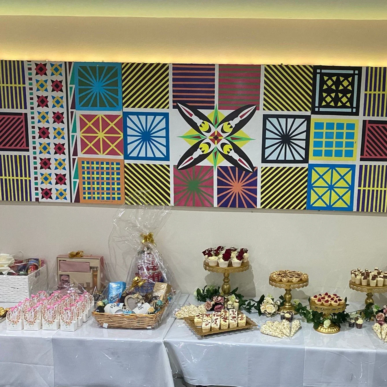 A decorative wall with geometric pattern artwork above a table set with a variety of desserts and gift baskets. The table is covered with a white cloth and features treats such as pastries, cupcakes, and wrapped gift items. The artwork consists of co