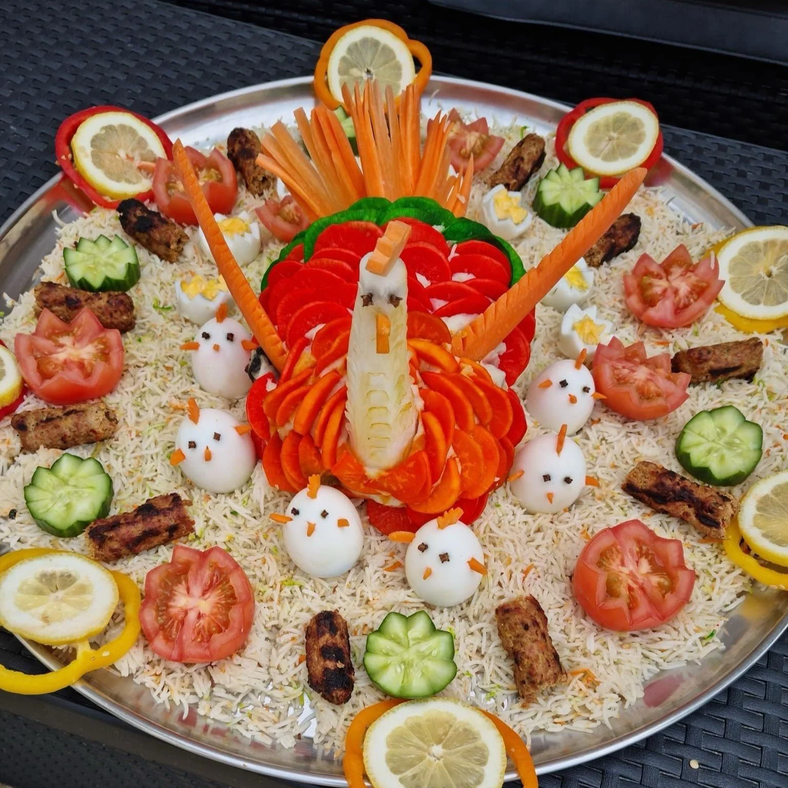 Decorative food platter with a rice base, garnished with carved vegetables and boiled eggs shaped like chicks. Centerpiece features carved vegetables resembling a peacock, surrounded by sliced cucumbers, tomatoes, lemon slices, and kebab pieces.
