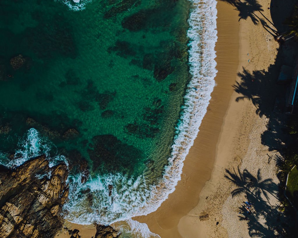 An aerial view of a sandy beach with clear turquoise water and shadows of palm trees