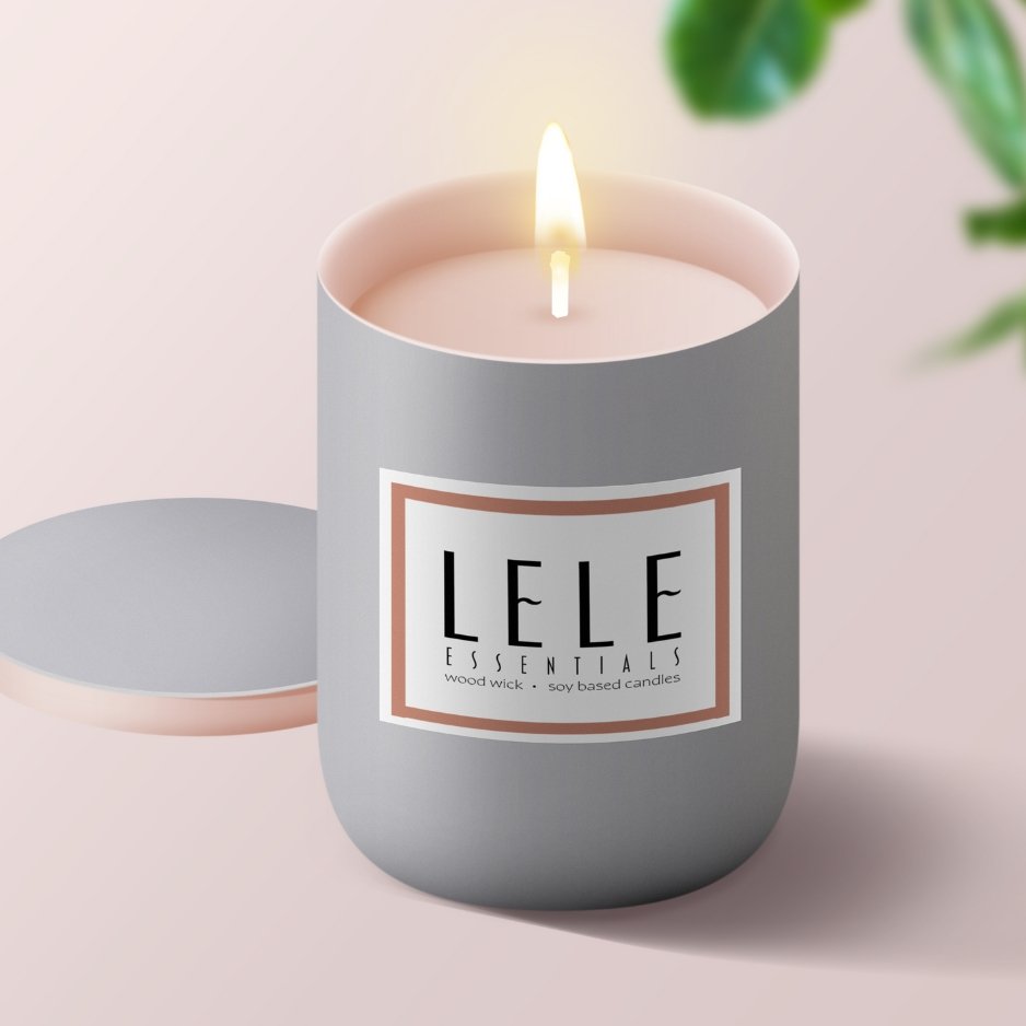A lit soy-based candle in a gray container with a label reading "LELE ESSENTIALS" and "wood wick," with green leaves in the background. Graphic Design Service by Capital Content.