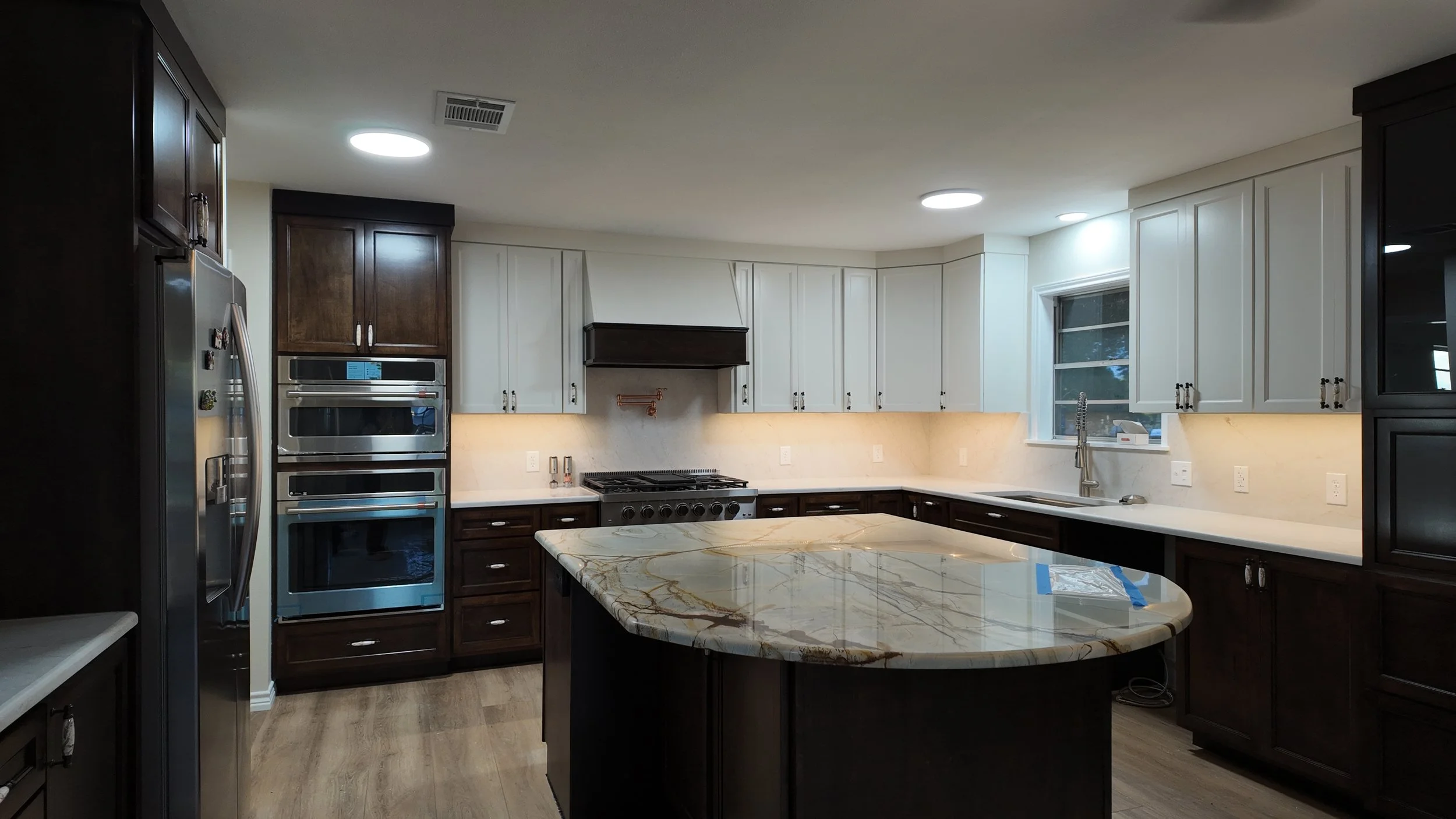 Modern kitchen with dark wood cabinets, white upper cabinets, stainless steel appliances, a marble island, a large window, and recessed ceiling lights.