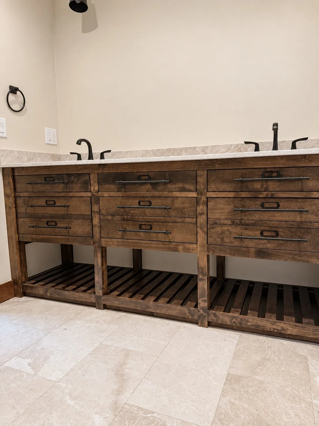 Dark wood double vanity with black bar pulls, pale countertops, two black faucets, and open slatted shelving.